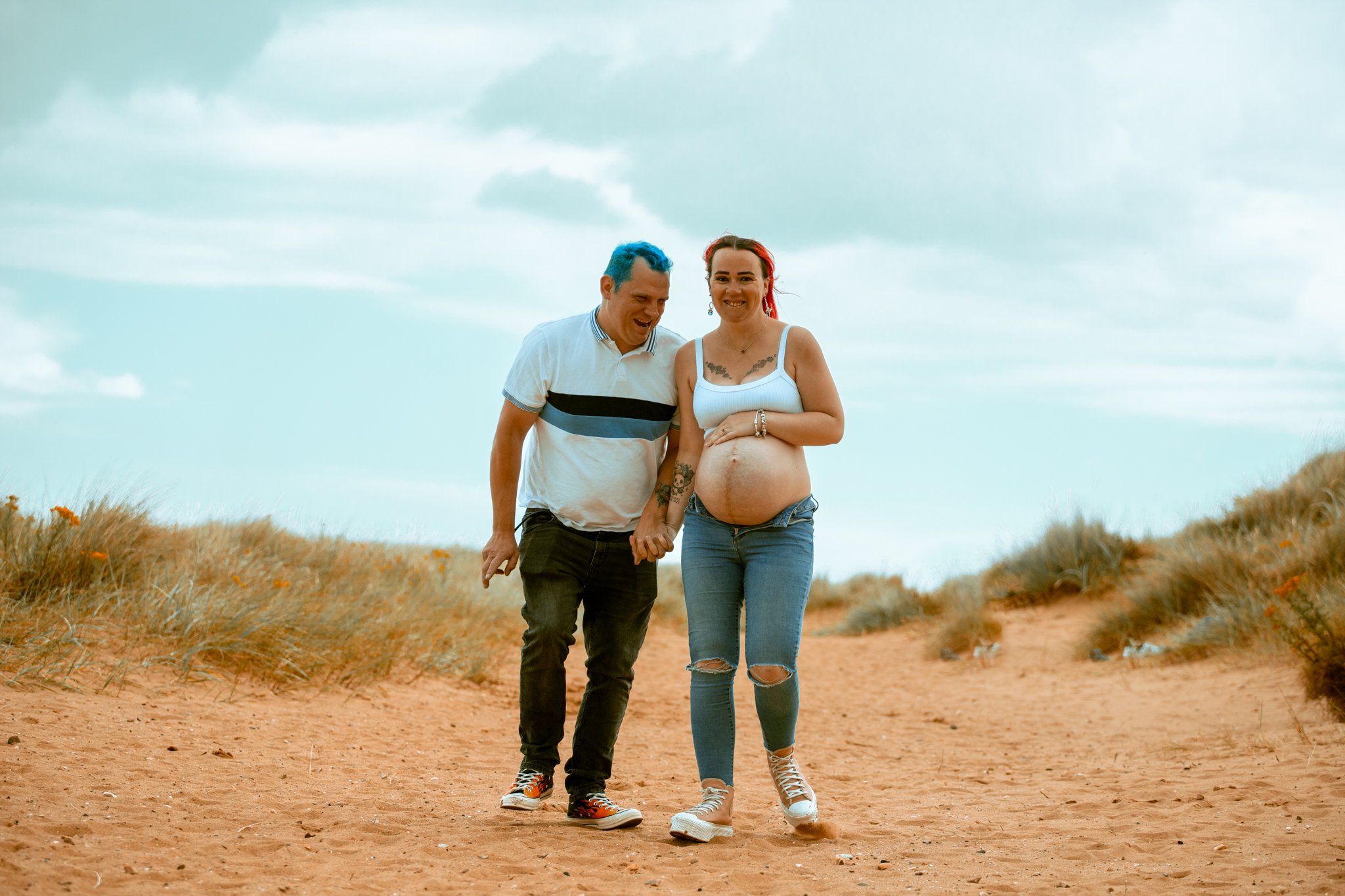 A pregnant woman and her partner walking hand-in-hand on a sandy trail in a desert landscape with dry grasses and a cloudy sky.