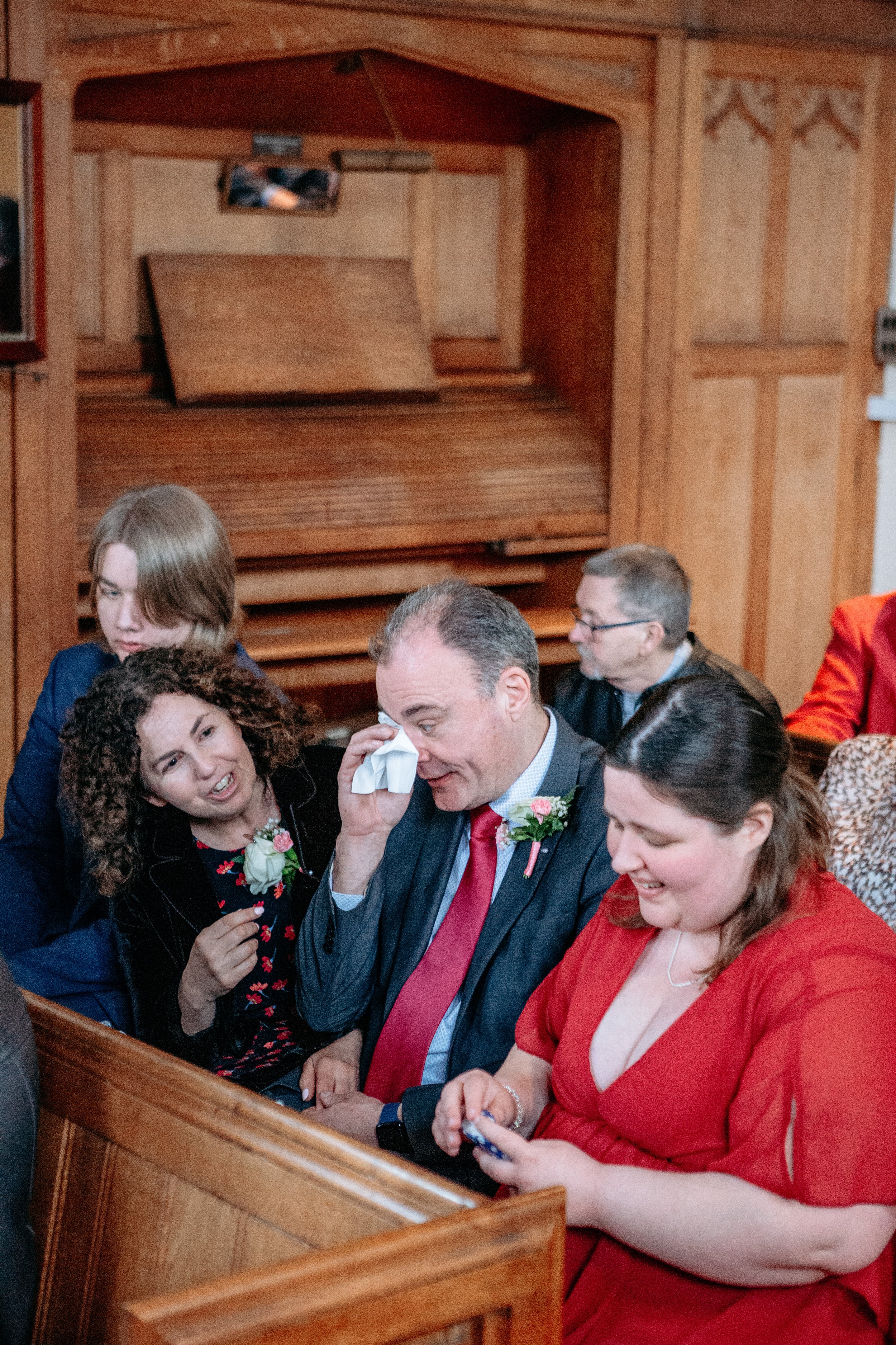Group of people dressed in formal attire, sitting in a wooden-paneled room during a wedding or celebration, with one man wiping his eye with a tissue, signifying an emotional moment.