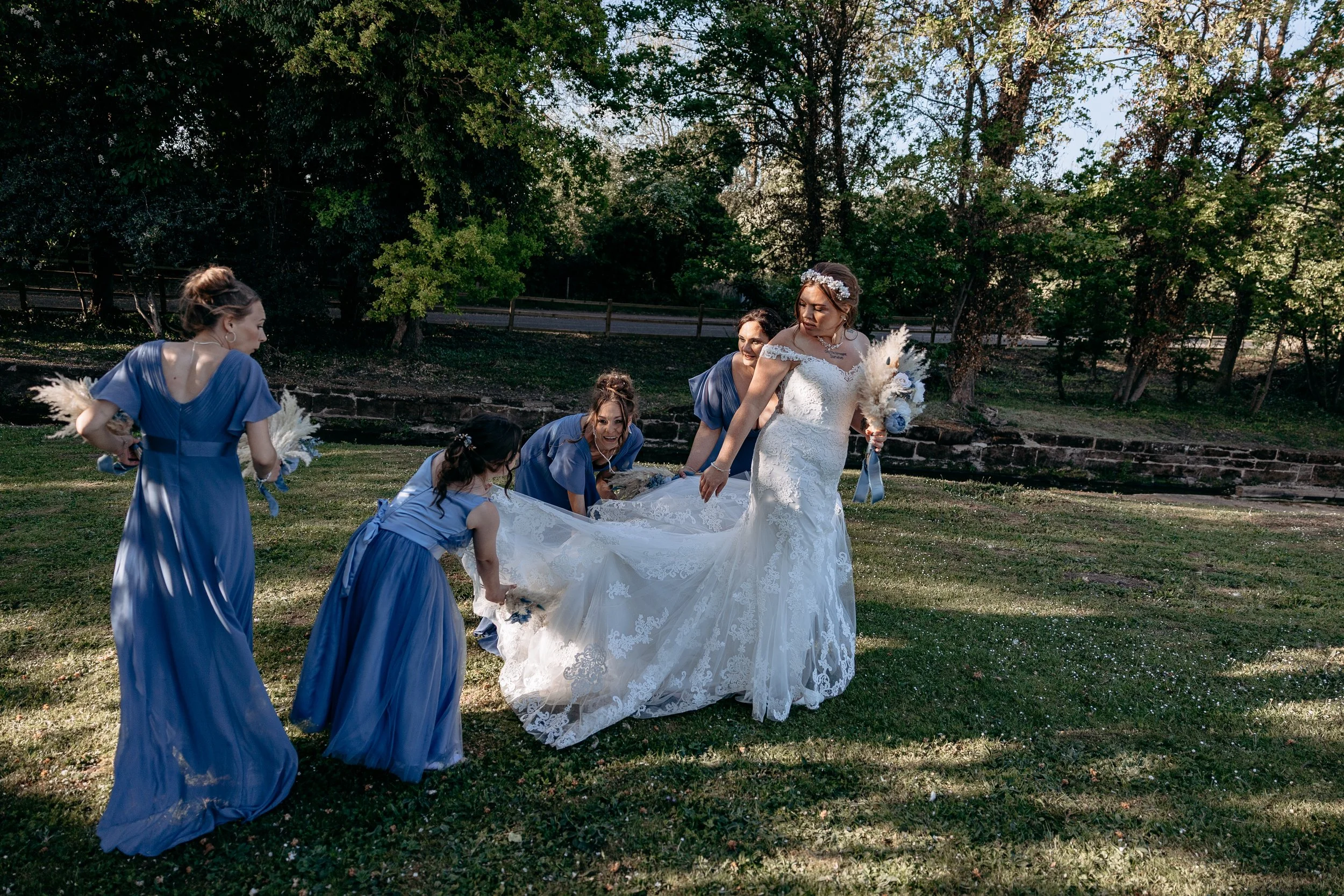 A bride in a white lace wedding dress and floral headband holds a bouquet of white and blue flowers, standing on a grassy area with bridesmaids in blue dresses around her, helping with her dress in a garden setting with trees in the background.