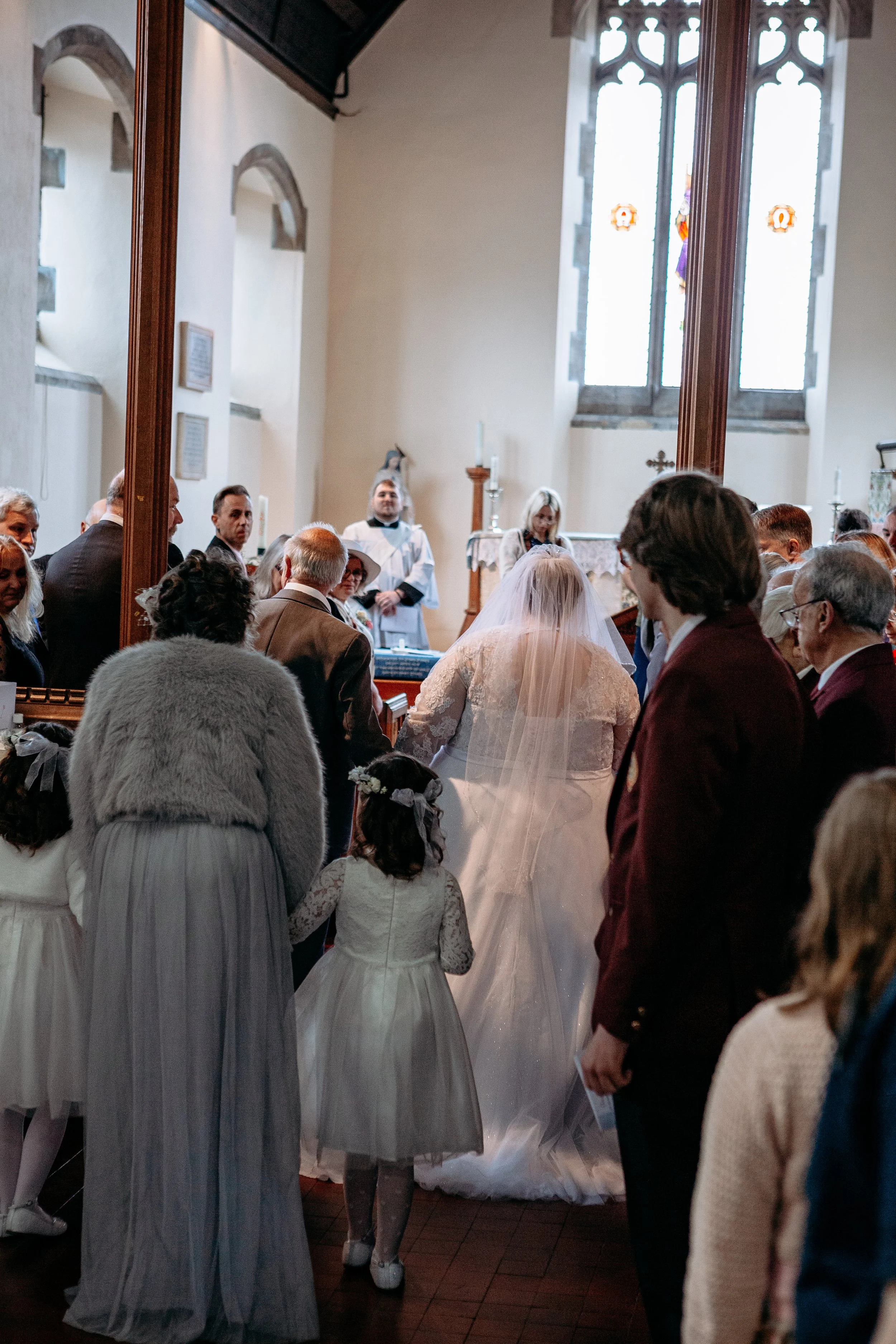 Bride in a white wedding dress and veil walking up the aisle in a church, surrounded by guests on both sides, during a wedding ceremony.