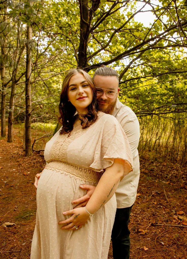 A pregnant woman and a man in a forest during daytime, with trees and green leaves in the background. The woman is wearing a beige dress, and the man is standing behind her, hugging her with both hands on her belly.