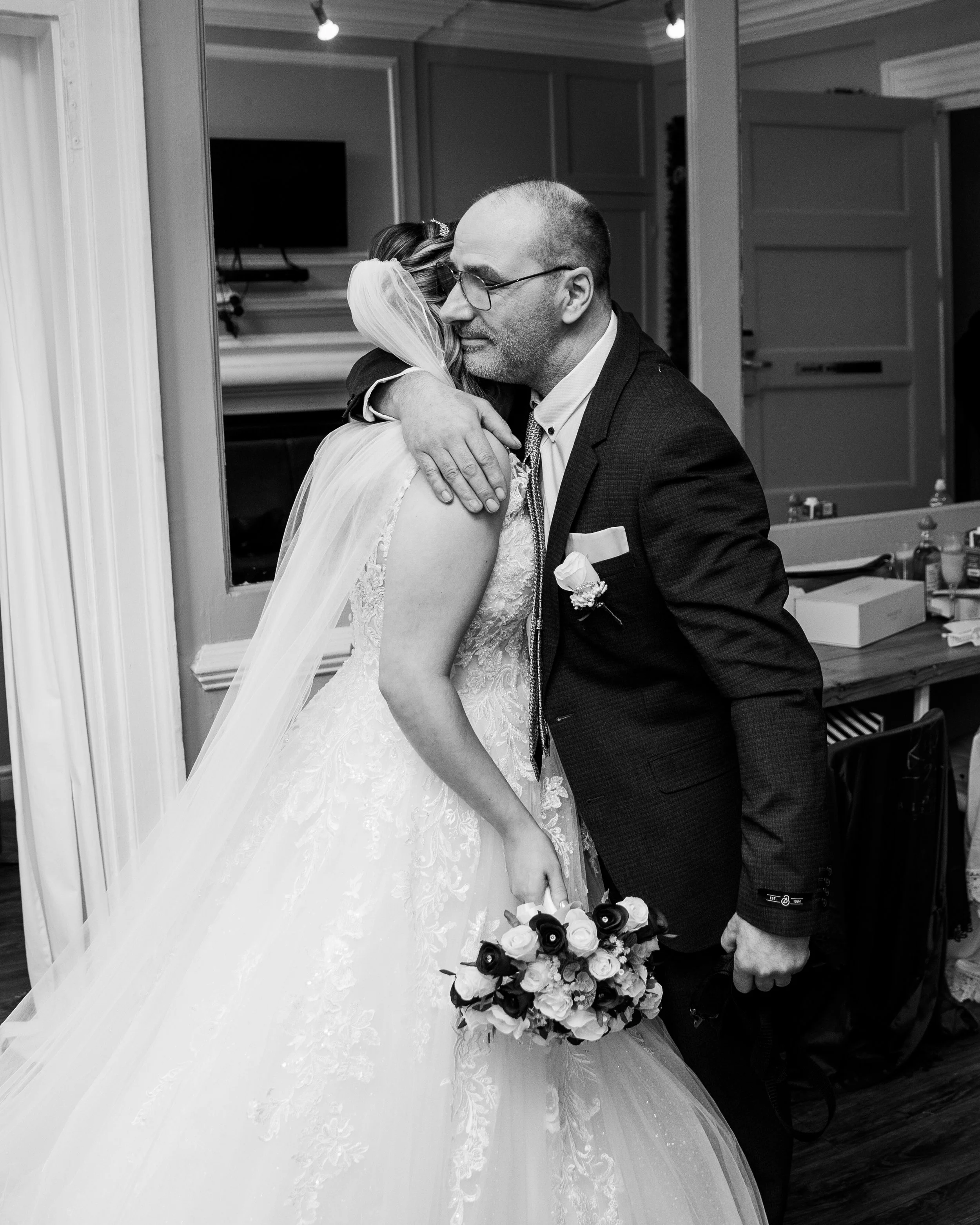 Black and white photo of a bride hugging a man, possibly her father, in a room. The bride wears a wedding dress with a long veil and holds a bouquet. The man wears a suit with a boutonniere and glasses.