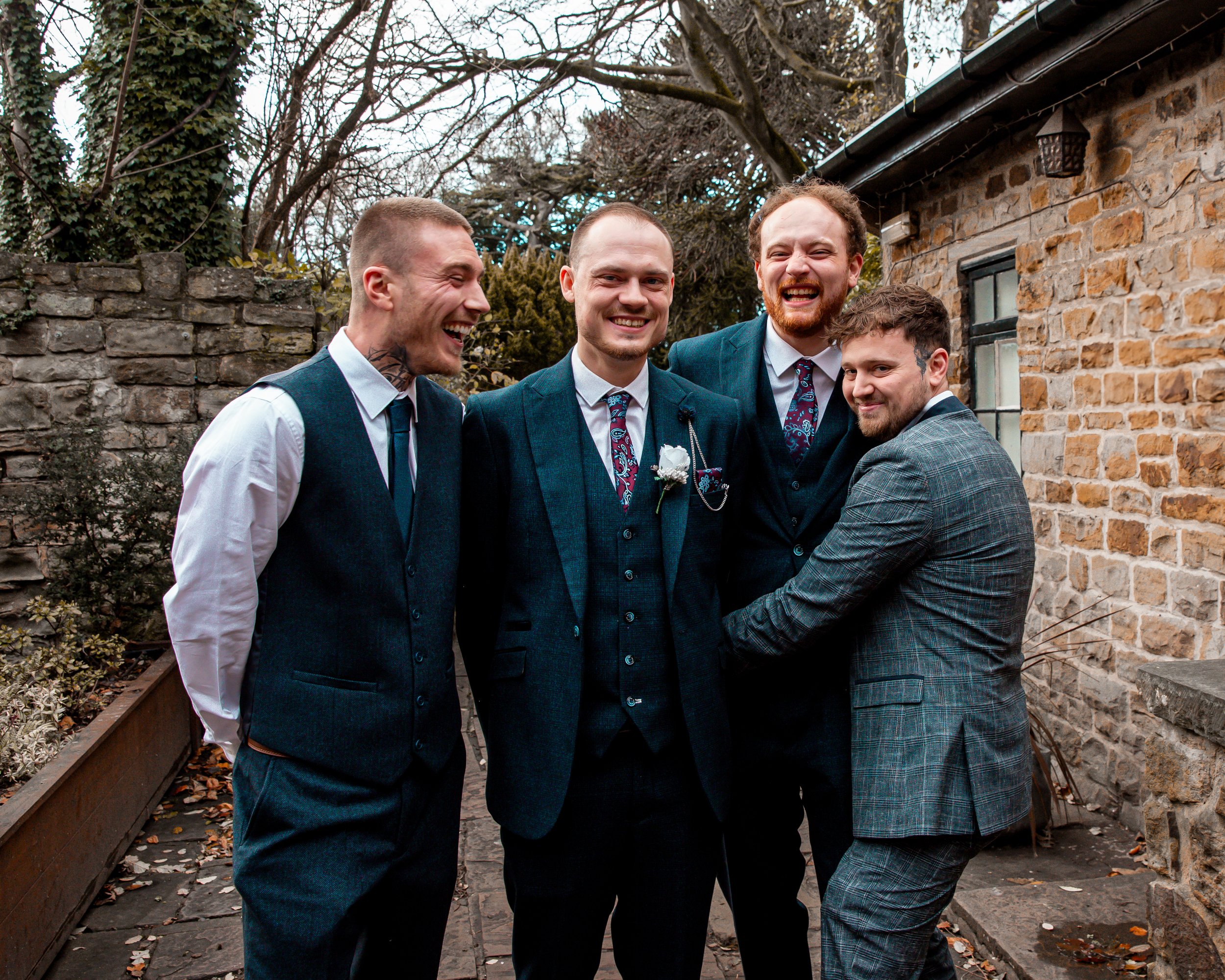 Five men in suits smiling and laughing together outdoors in a courtyard with stone walls, trees, and cloudy sky.