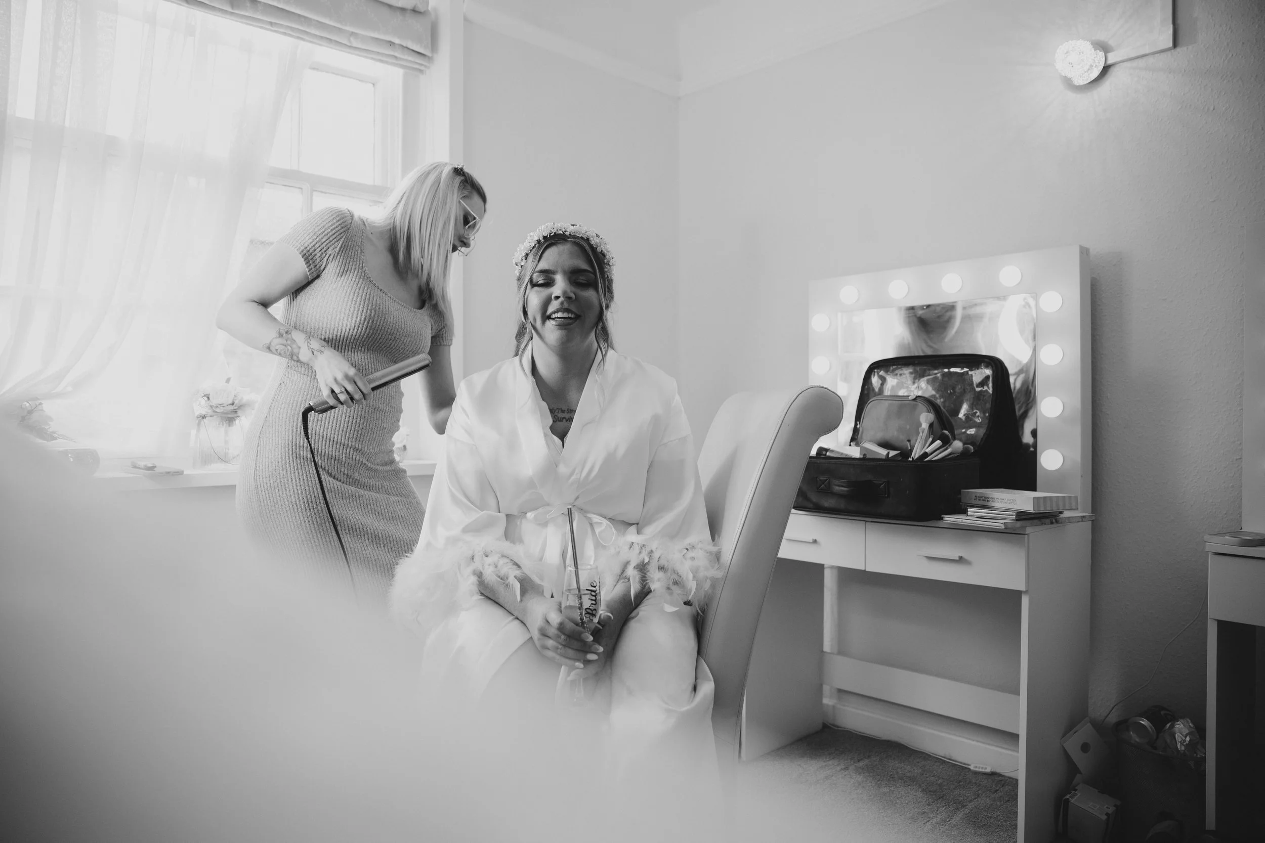 A woman in a white dressing gown sitting in a makeup chair, smiling, while another woman styles her hair with curling iron in a room with a mirror surrounded by lights.