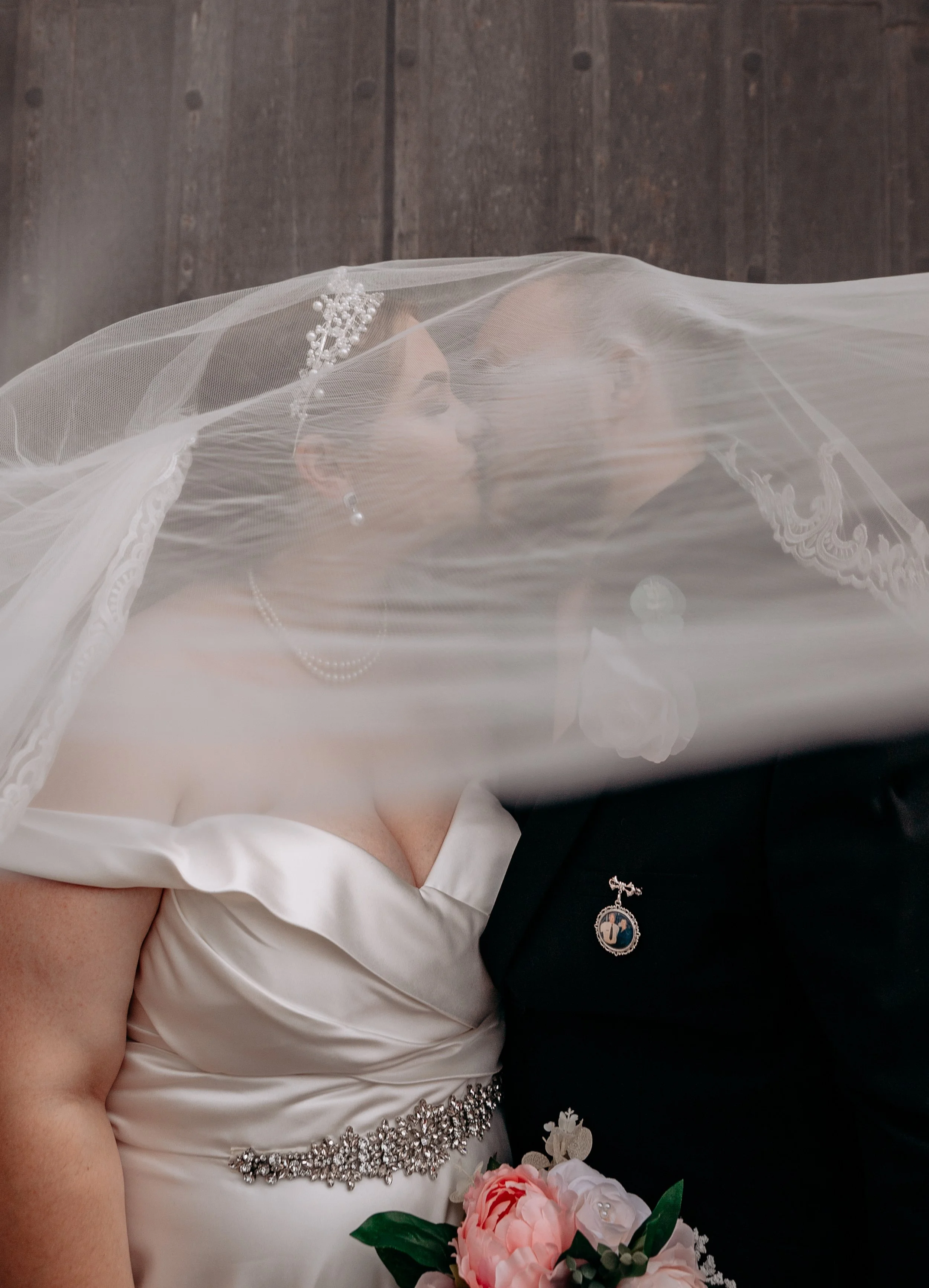 A bride and groom sharing a kiss behind a veil during wedding ceremony, with the bride wearing a satin gown with beaded waist detail and pearl jewelry, and the groom in a black suit with a lapel pin. The bride is holding a bouquet of pink and white f