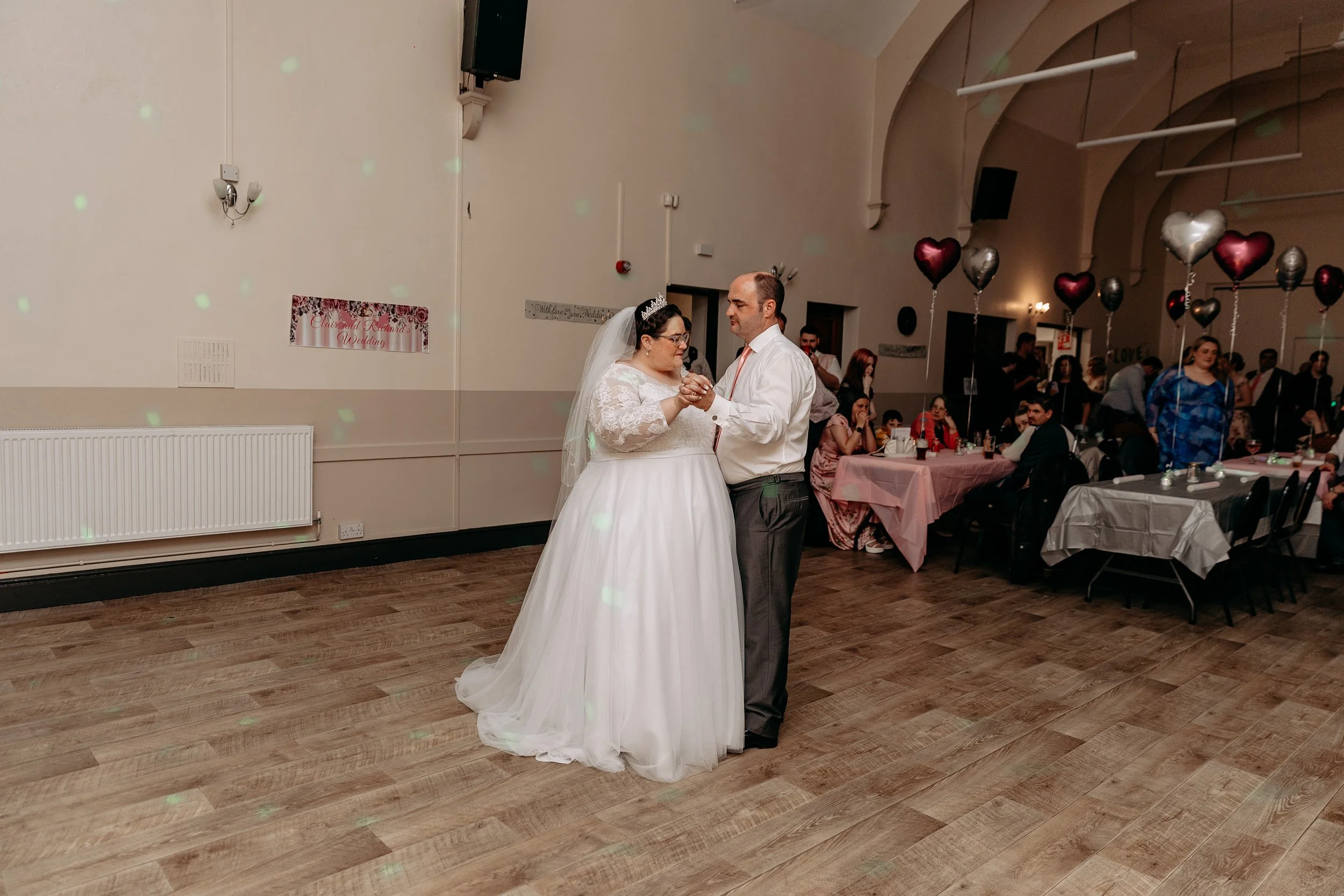 Bride and groom dancing at their wedding reception, with guests seated at decorated tables in the background and heart-shaped balloons.