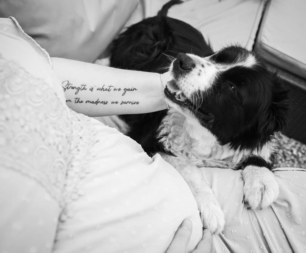 A person with a tattoo on their arm sitting on a bed, smiling while hugging a happy black and white dog. The tattoo reads 'Strength is what we gain from the madness we survive.'