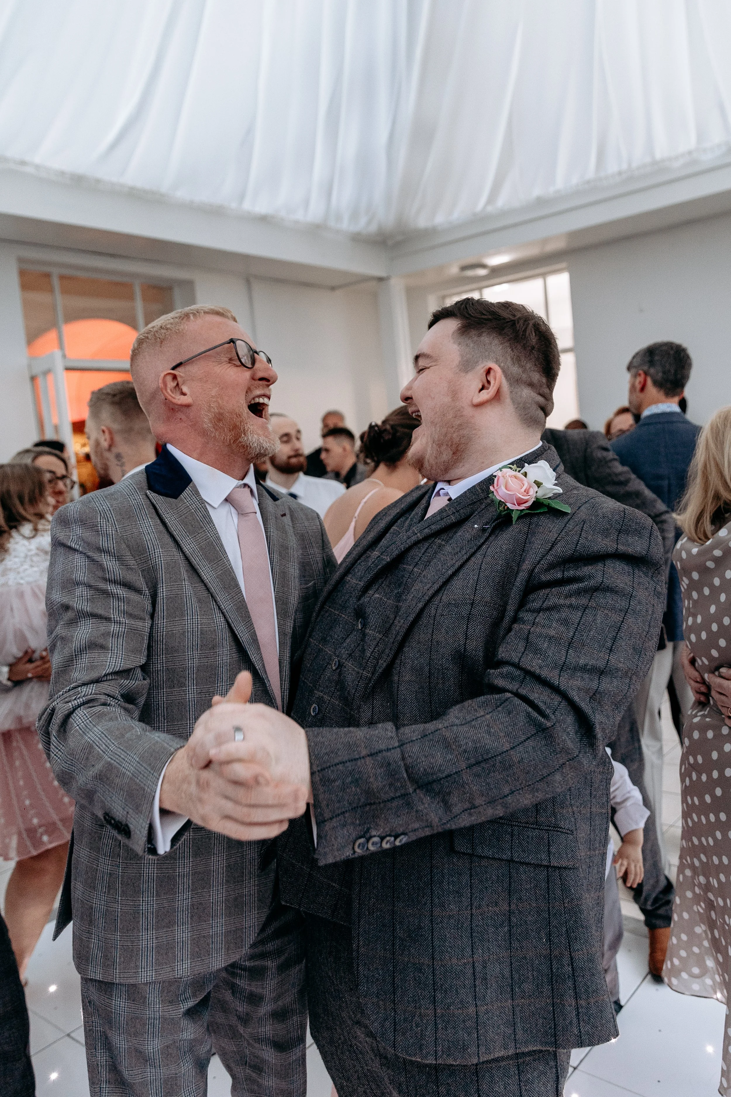 Two men are dancing and laughing together at a wedding celebration, dressed in suits with pink ties, surrounded by guests in a decorated indoor venue.