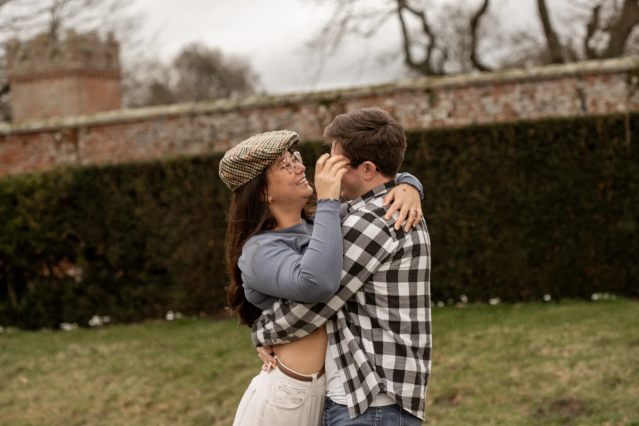 A young couple embracing outdoors on a cloudy day, smiling at each other, with a brick wall and hedge in the background.