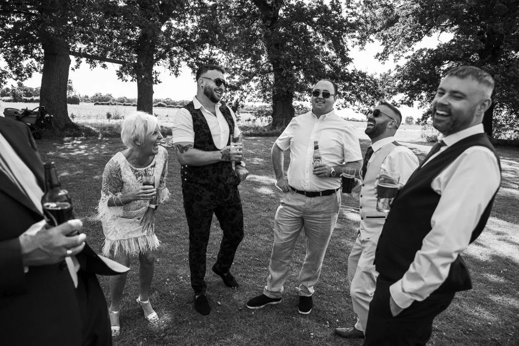 A group of six adults outdoors, laughing and holding drinks, standing under trees in a grassy area.