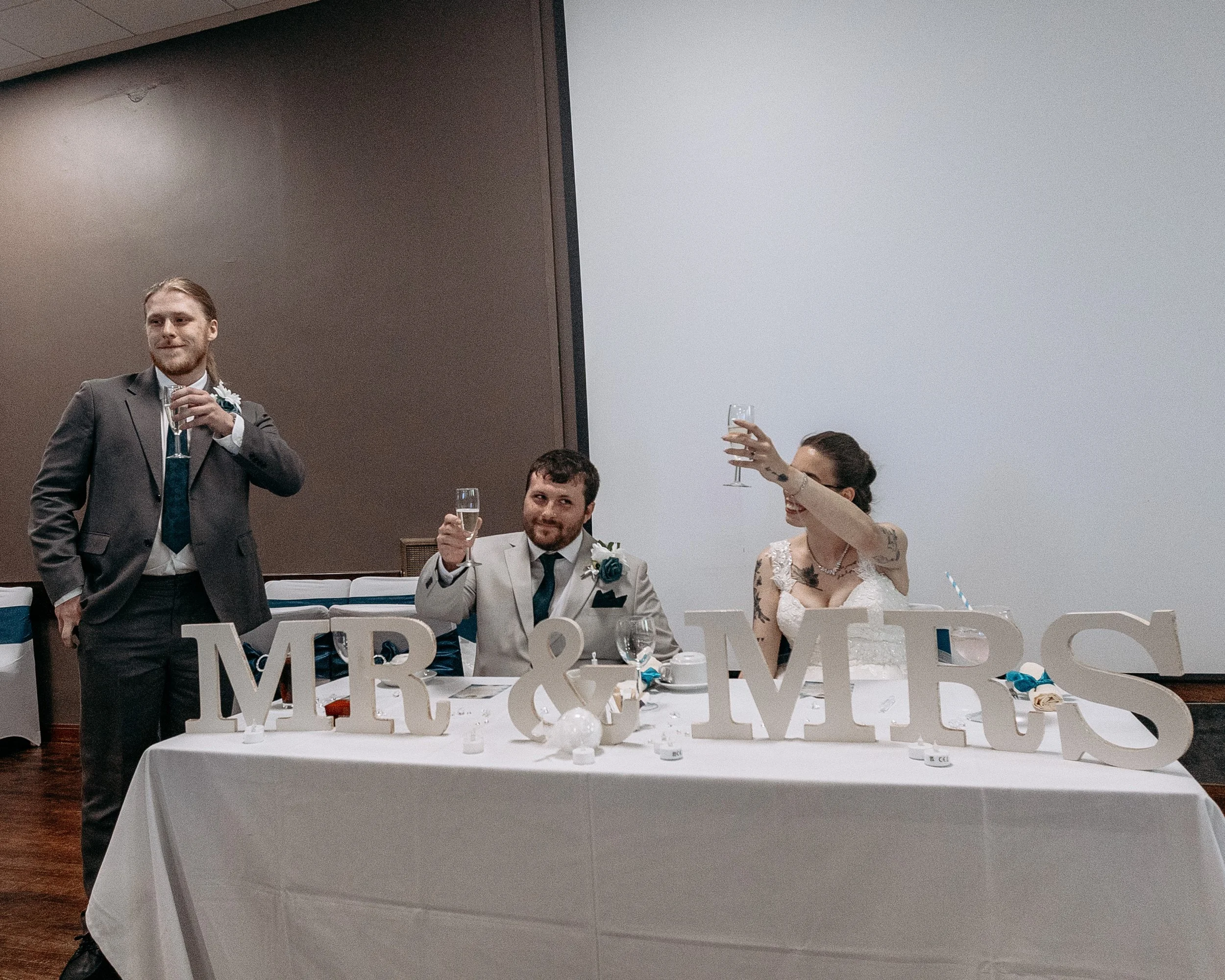 A wedding celebration with the bride, groom, and a guest raising glasses in a toast at the head table decorated with large 'MRS & MR S' signs.