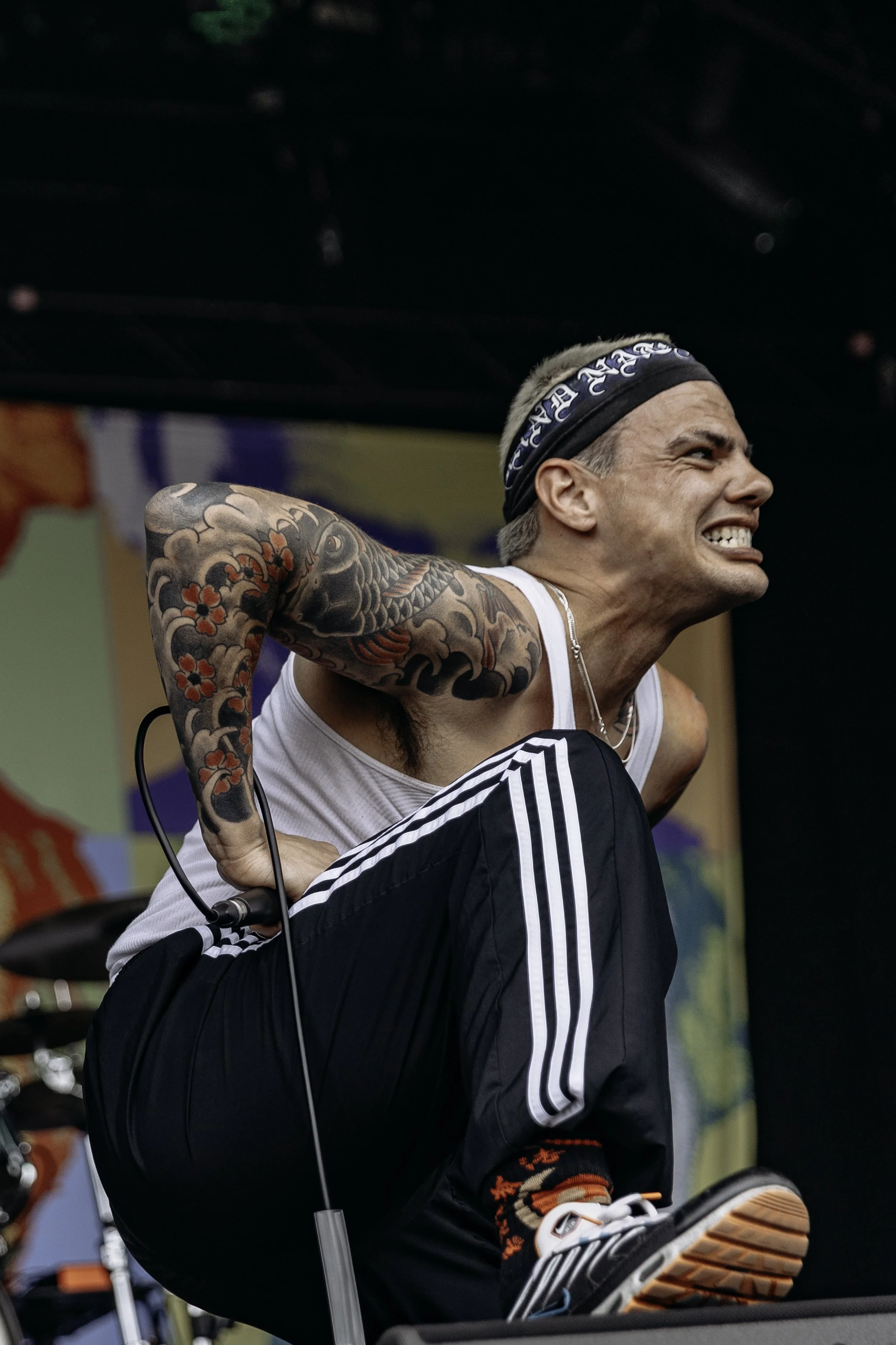 A man with a band_headband, tattoos on his arm, wearing a white tank top and black Adidas pants, is performing on stage, kneeling with an intense expression.