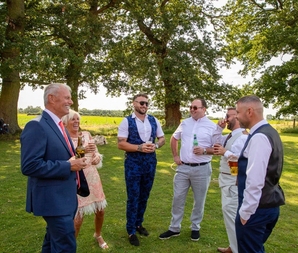 Group of people socializing outdoors under large trees on a sunny day, holding drinks and smiling.
