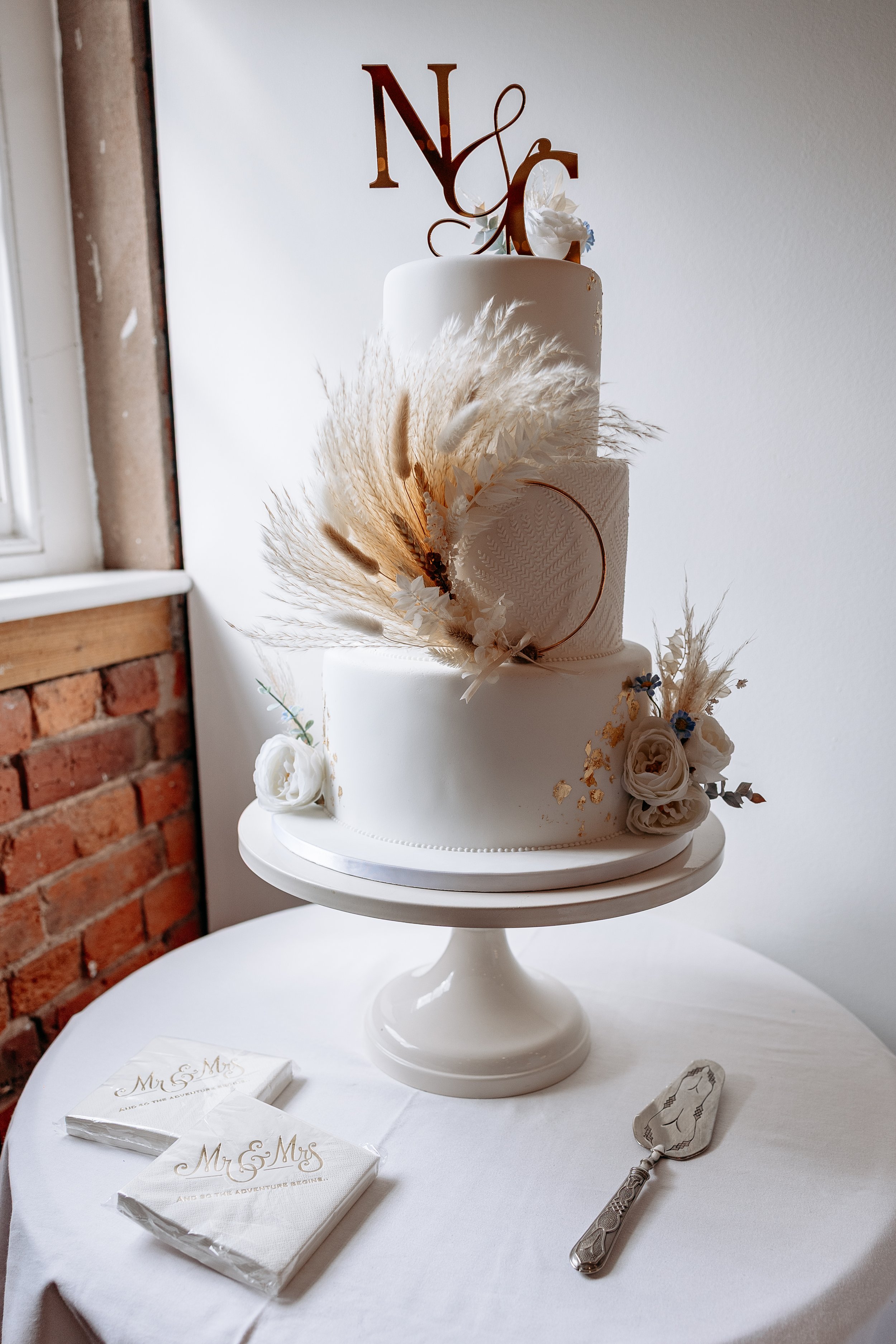 A three-tier white wedding cake decorated with dried flowers and gold accents, with a golden cake topper featuring the letter N and the letter C intertwined, placed on a white cake stand on a round table with a white tablecloth. There are two napkins