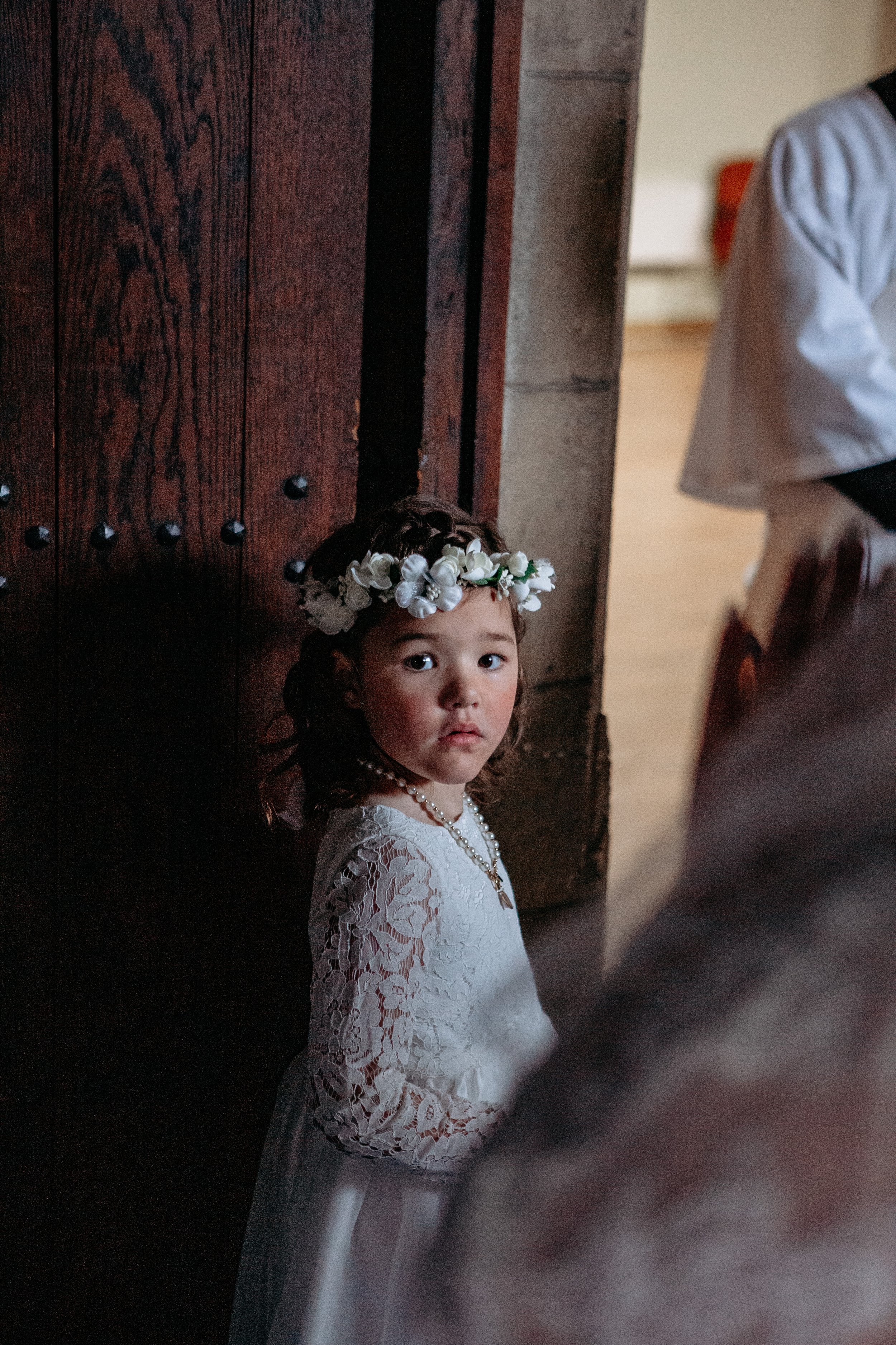 A young girl with a flower crown and pearl necklace looking towards the camera in a church or chapel setting.