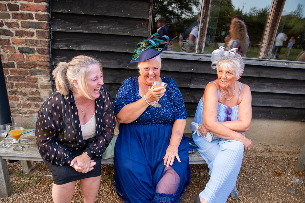 Three women sitting on a wooden bench outdoors, laughing and enjoying drinks at a social gathering, with people in the background reflected in the window.