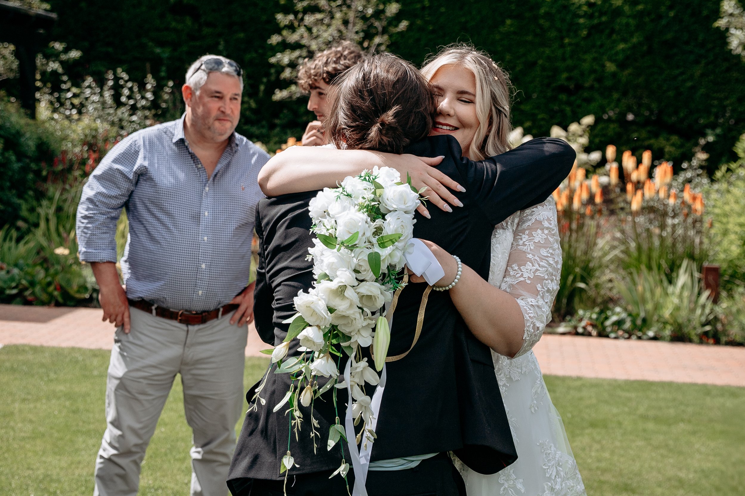 Bride hugging a person in a garden setting during a wedding ceremony, with two other people standing nearby.