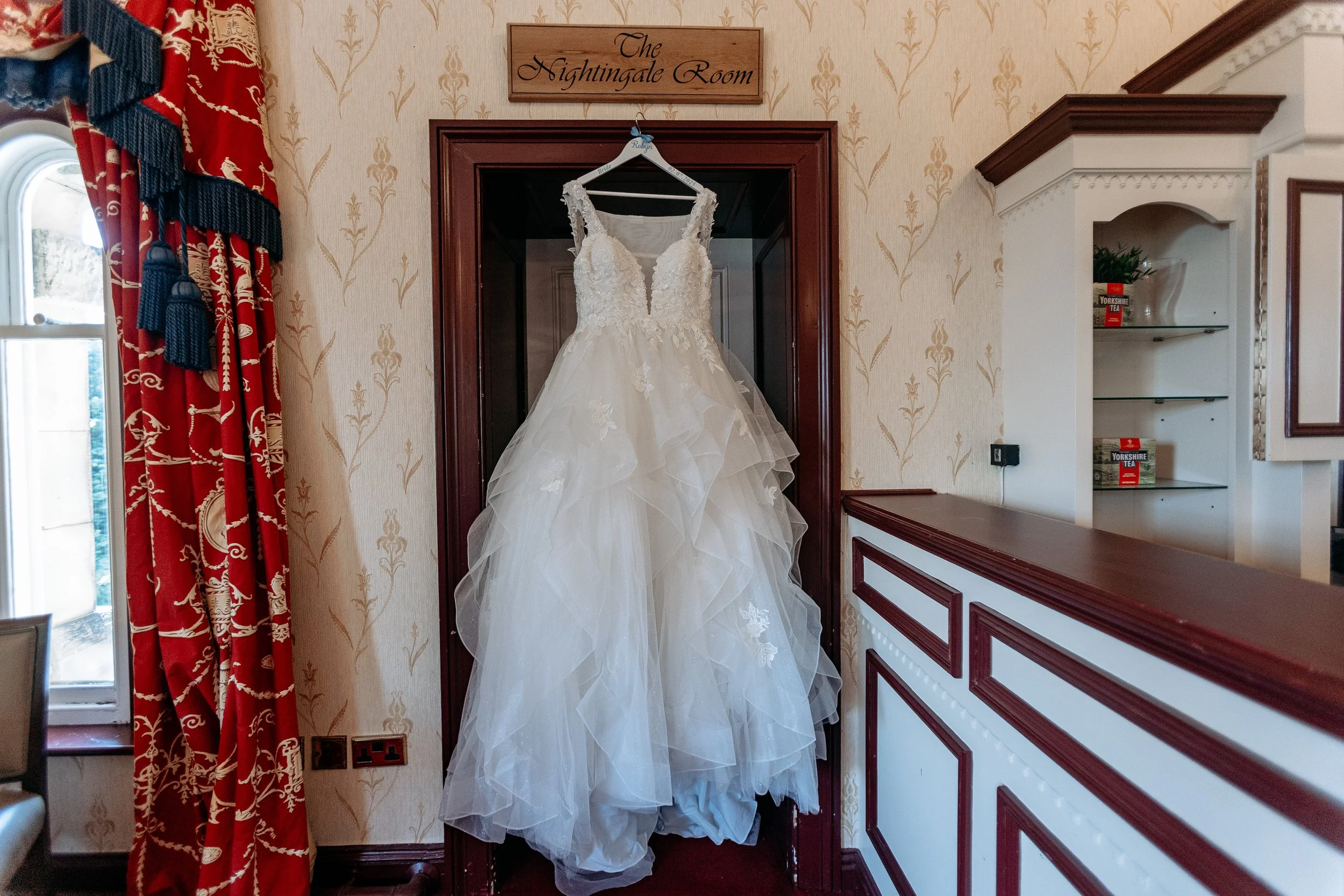 A wedding dress hanging on a hanger in a wooden closet nook labeled 'The Nightingale Room,' with red and gold curtains, a window, and a white cabinet with books and tea boxes visible in the background.
