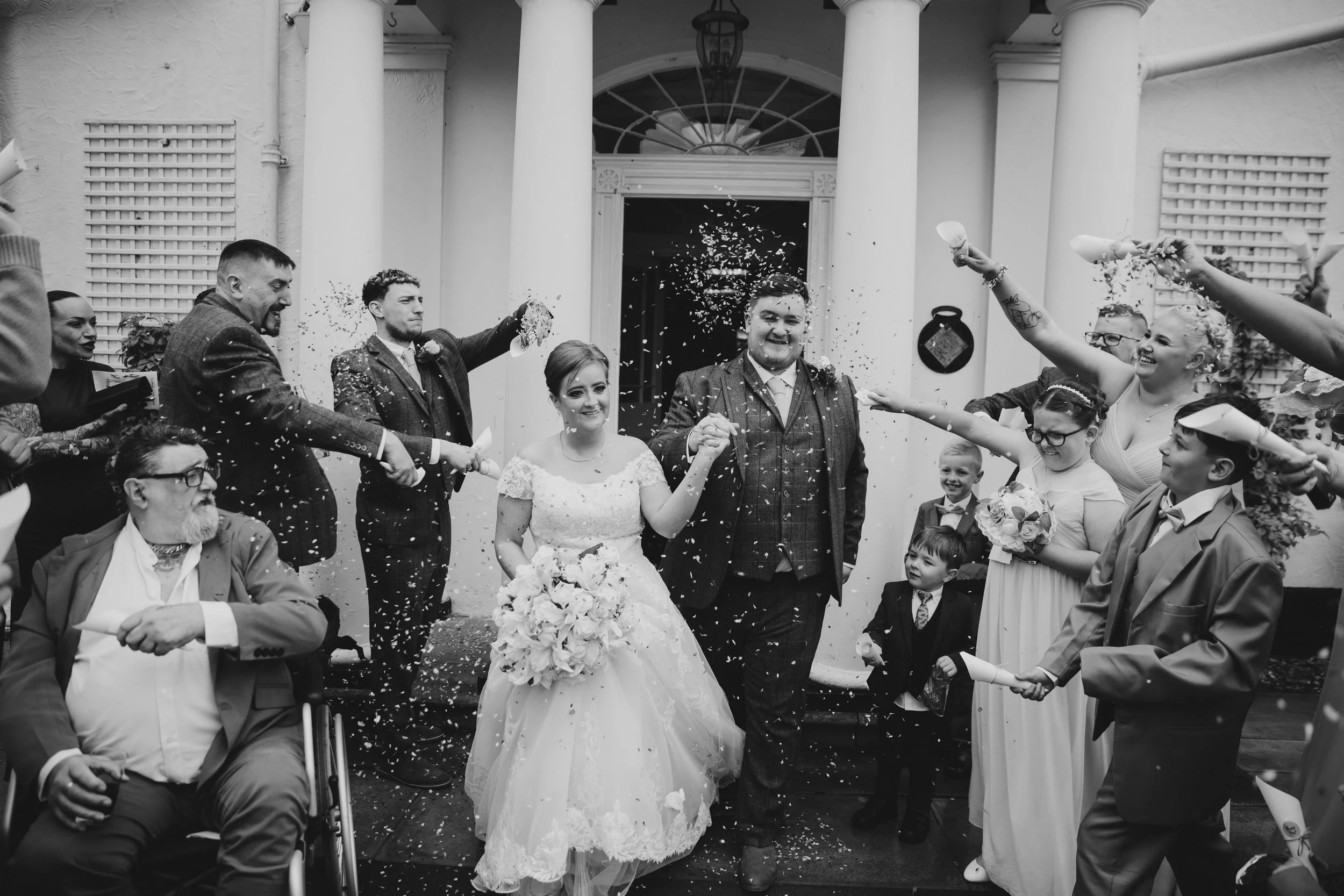 Black and white photo of a wedding celebration with a bride and groom walking out of a building, surrounded by guests throwing confetti. The bride is holding a bouquet and wearing a lace wedding dress, while the groom wears a suit. The guests include