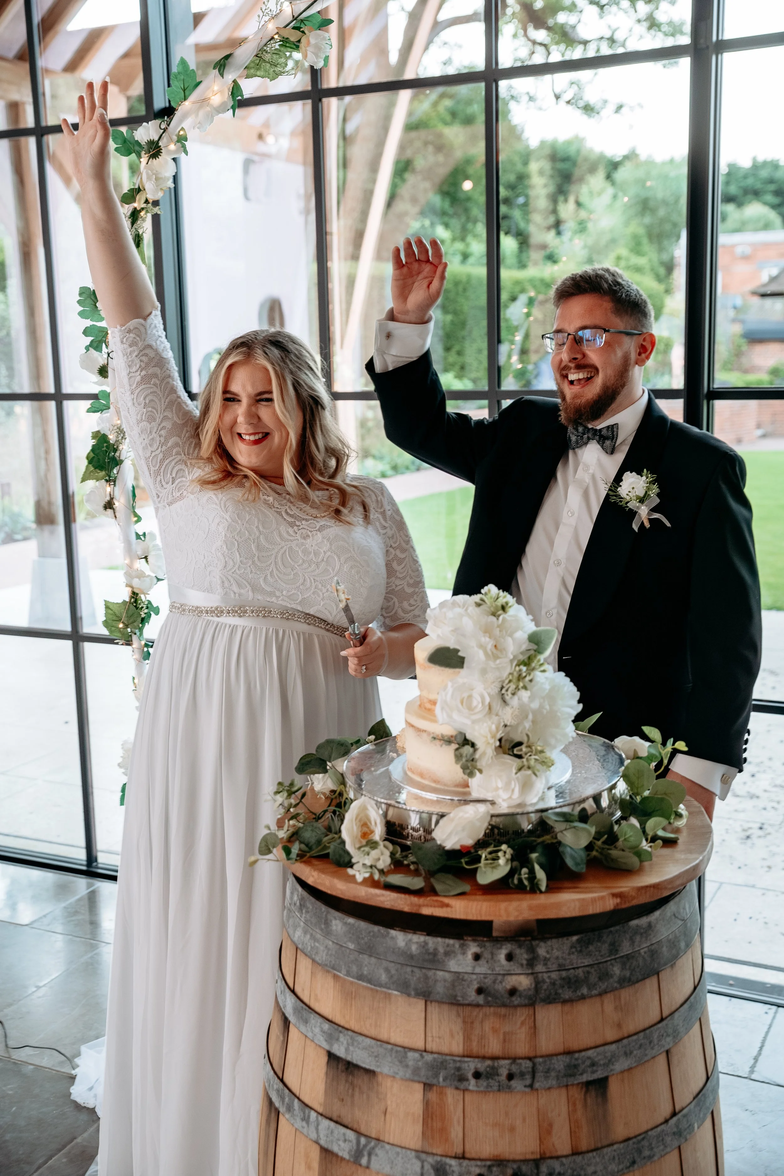 A newlywed couple celebrating their wedding with a cake cutting ceremony. The bride is in a white lace gown, and the groom is in a black tuxedo. They are smiling and raising their hands. The wedding cake is on a wooden barrel decorated with white flo