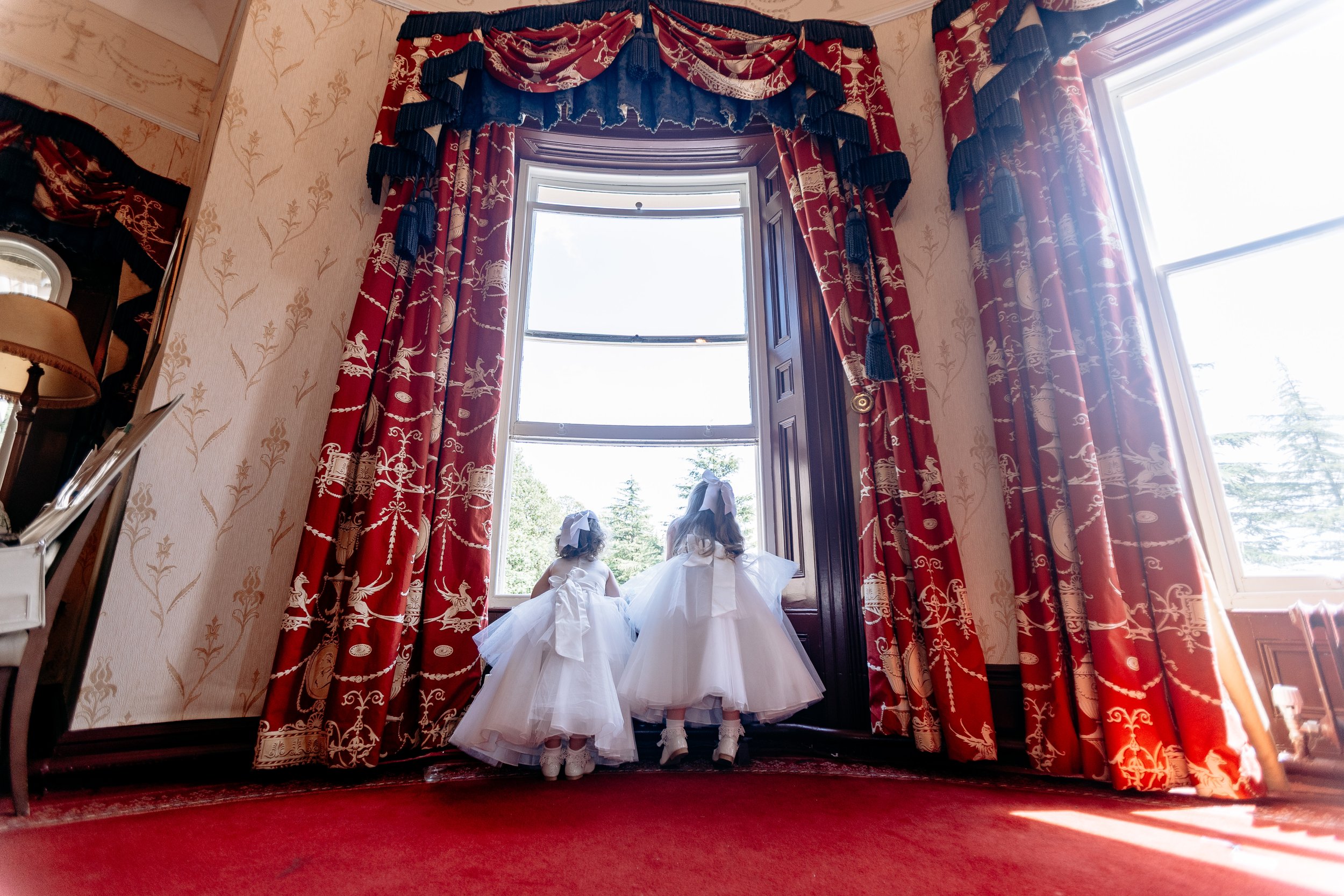 Two young girls in white dresses and headbands are standing at a large bay window, looking outside on a bright day. The room features red patterned curtains, floral wallpaper, and a red carpet.