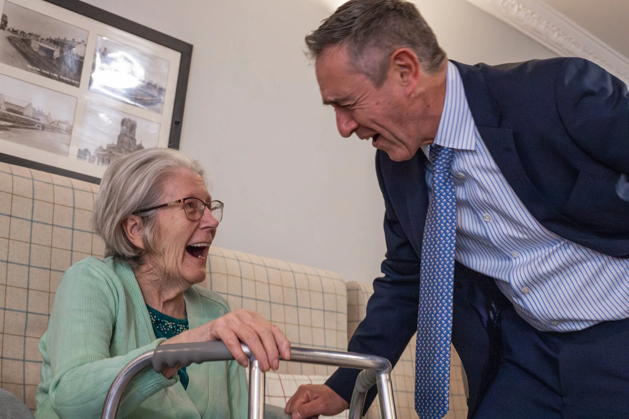 An elderly woman in glasses laughing with her mouth open, sitting on a sofa with a walker, interacts with a man in a suit and tie who is laughing and leaning towards her.