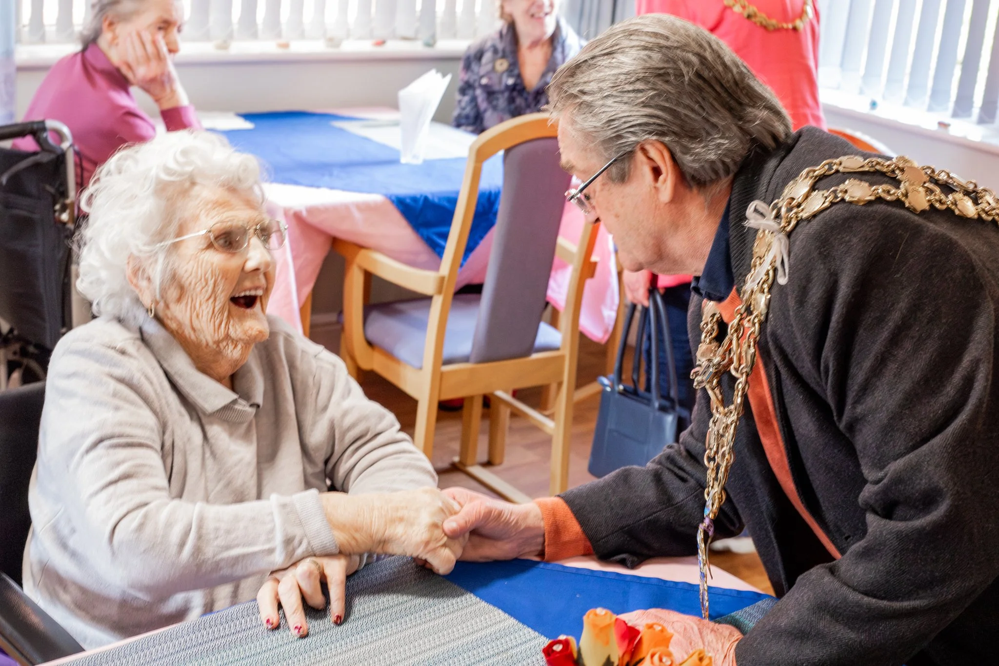 An elderly woman with glasses and white hair smiling as she shakes hands with a man wearing glasses and a chain tradition, at a social gathering.