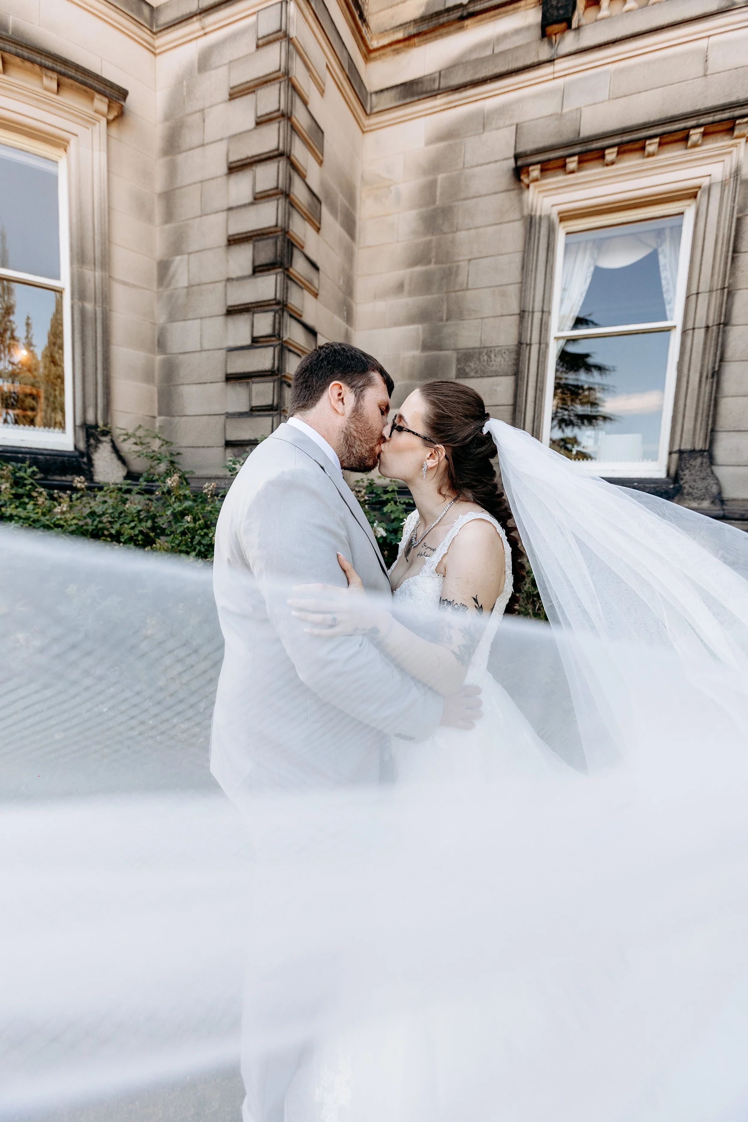 A newlywed couple kissing outdoors in front of an old stone building, with the bride wearing a white wedding dress and veil, and the groom in a light gray suit.