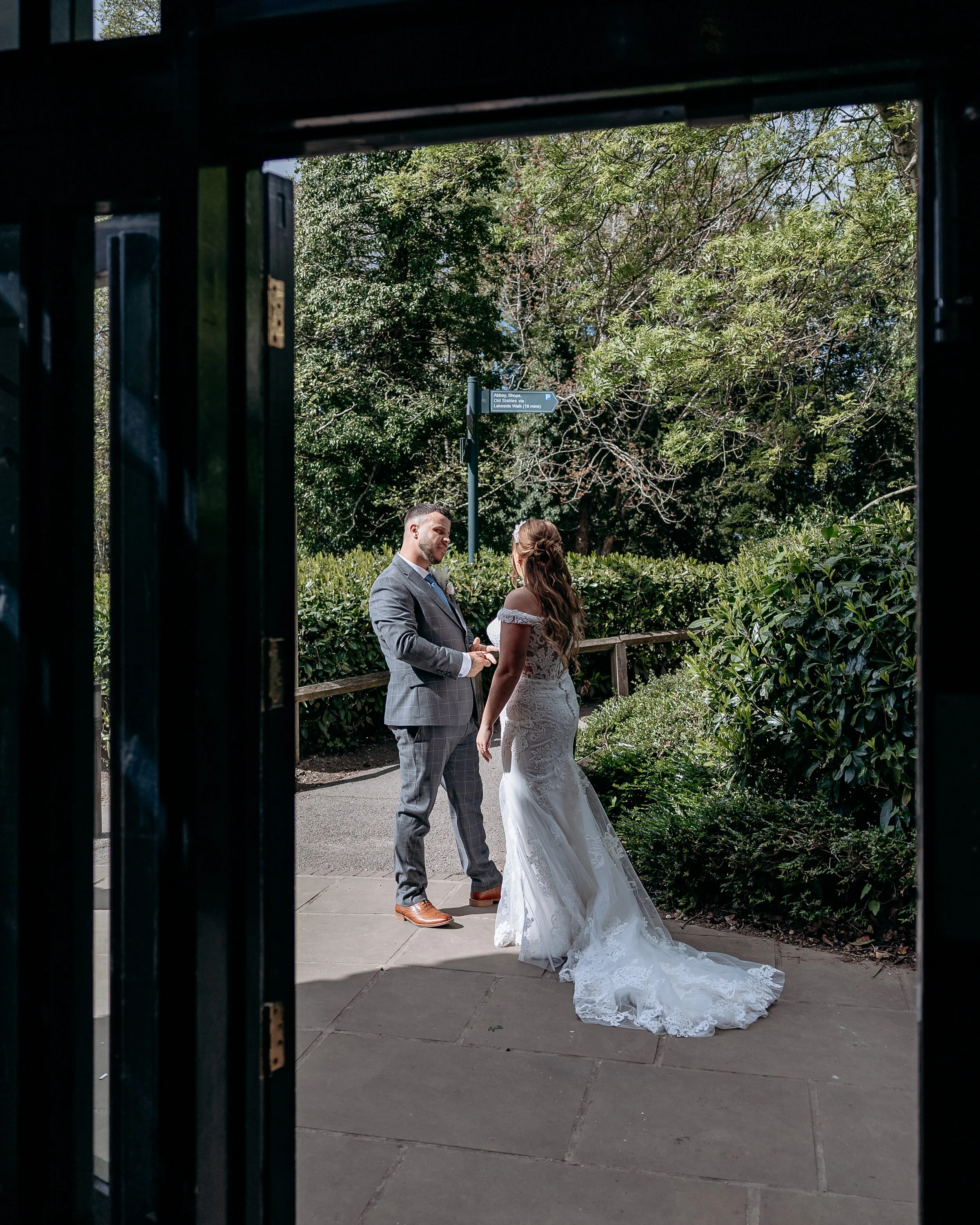A bride and groom are exchanging vows outdoors, seen through an open window, with greenery and trees in the background.
