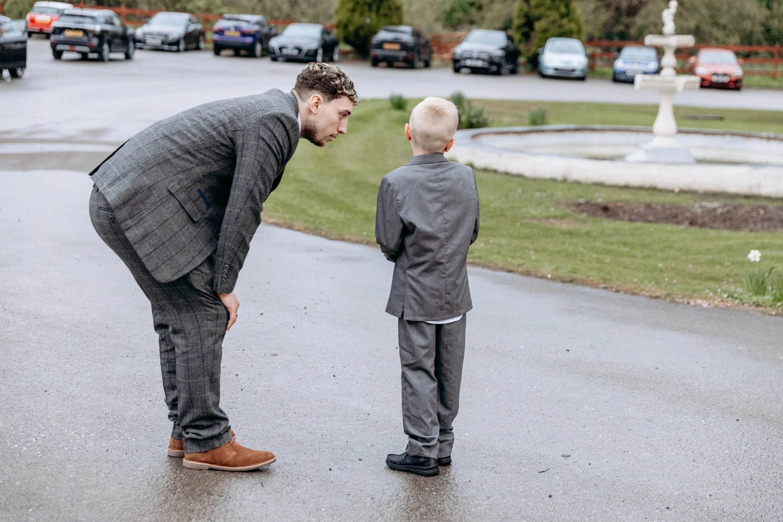 A man and a young boy in suits are standing on a wet pavement, having a conversation. The man is leaning forward, talking to the boy who has his back turned. A fountain, grass, and parked cars are visible in the background.