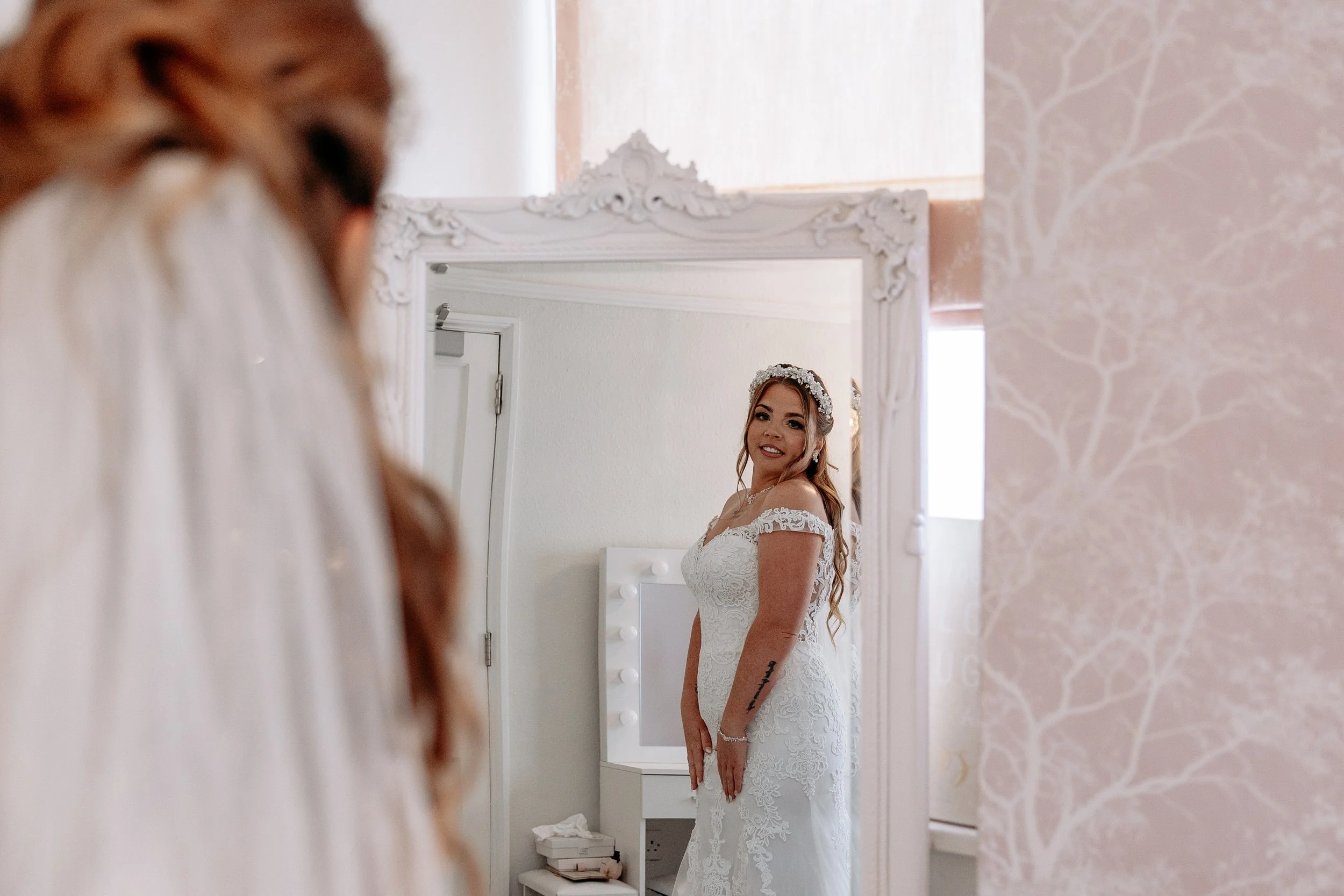A bride looking at herself in a mirror on her wedding day, wearing a white lace wedding dress and a floral headband.