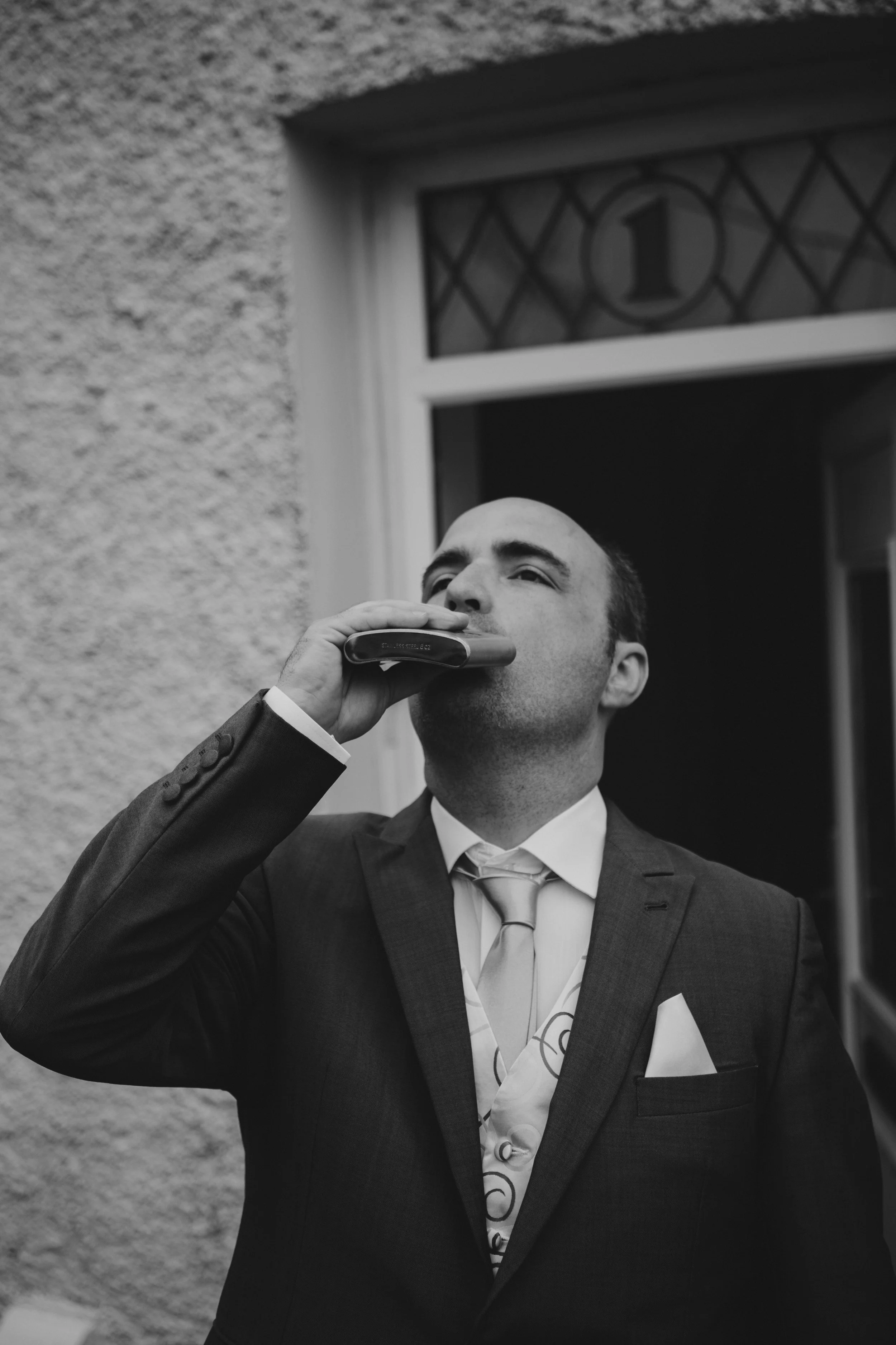 Man in business suit playing a harmonica on a porch outside a building with a window marked '1'.
