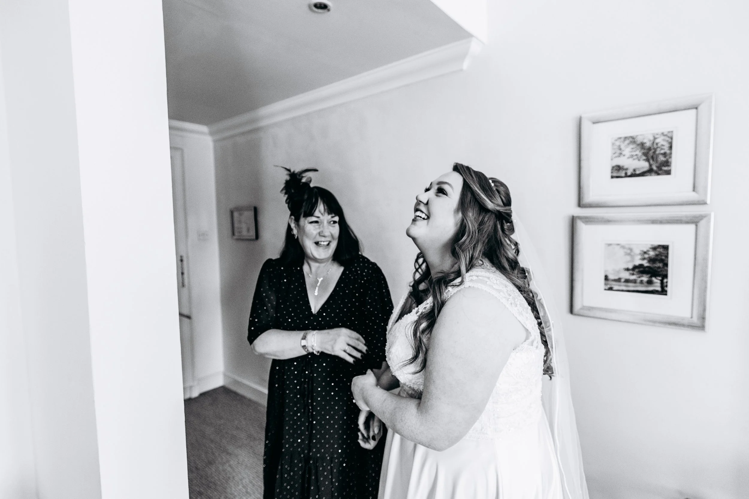 A woman in a wedding dress laughing with a woman in a polka dot dress, both appearing joyful, in a home interior with framed pictures on the wall.