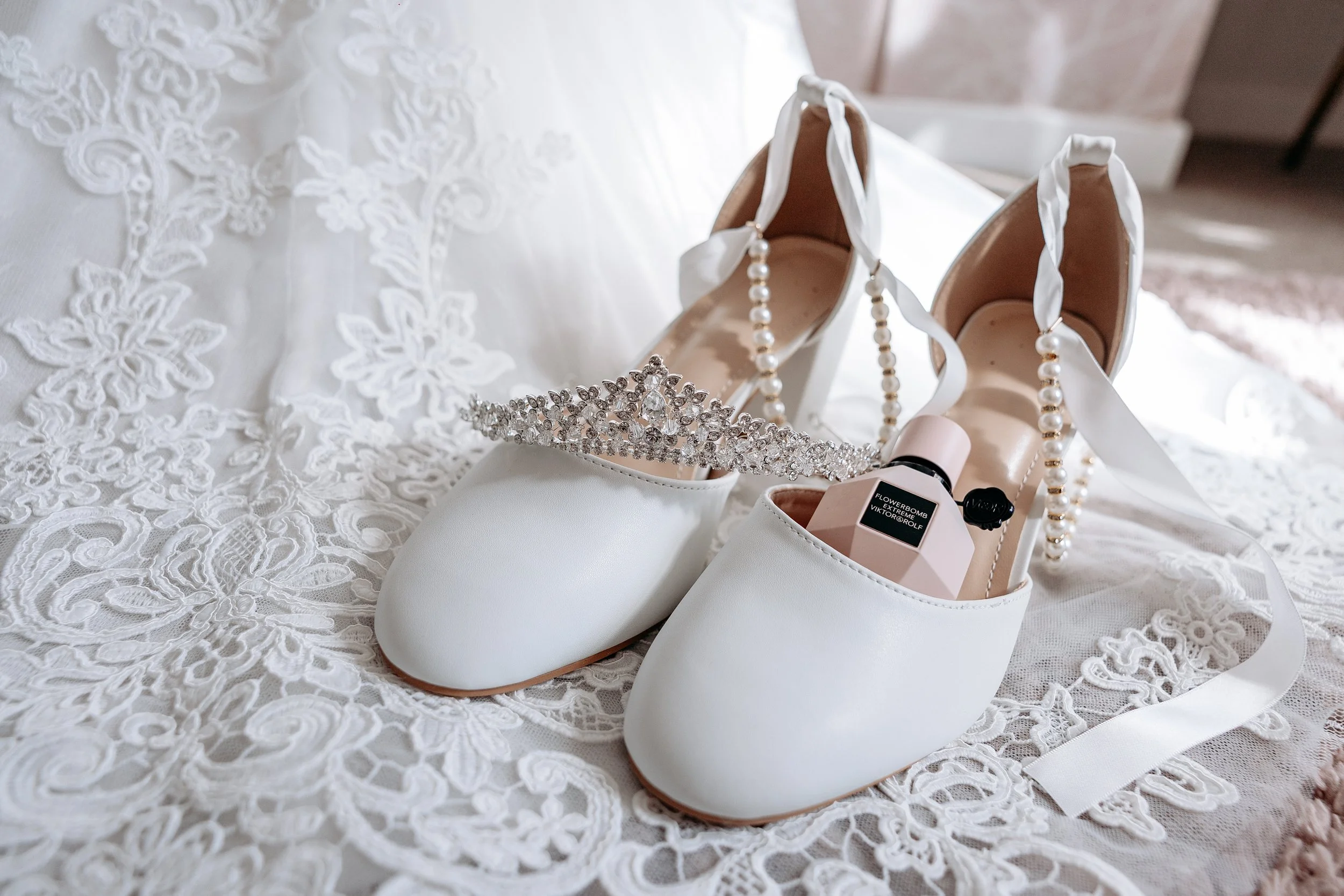 A pair of white bridal shoes with a pearl ankle strap, a rhinestone tiara, a bridal perfume, and a container of perfume, all placed on lace fabric.