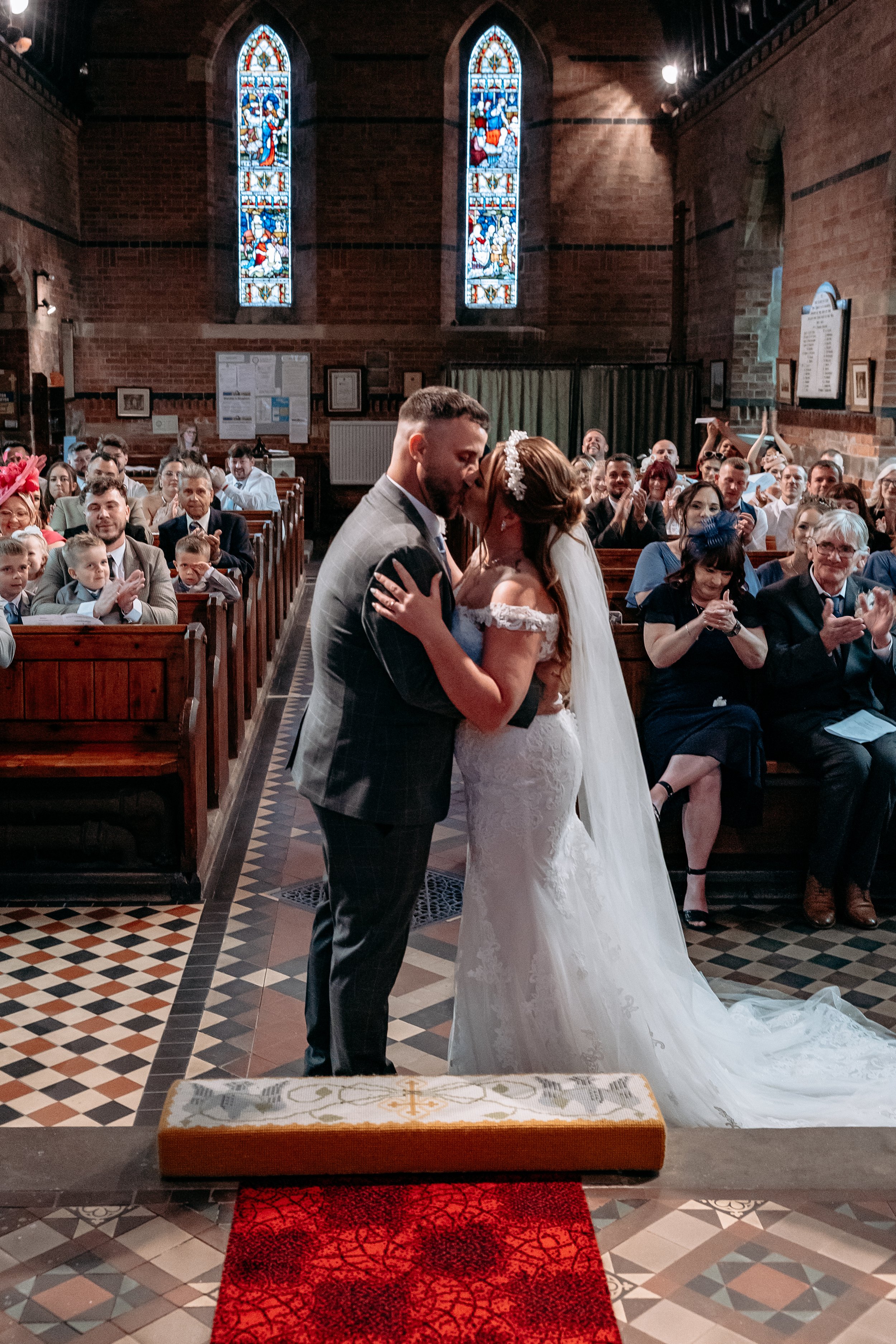 Bride and groom sharing a kiss during their wedding ceremony in a church, with guests clapping and smiling in the pews behind them.