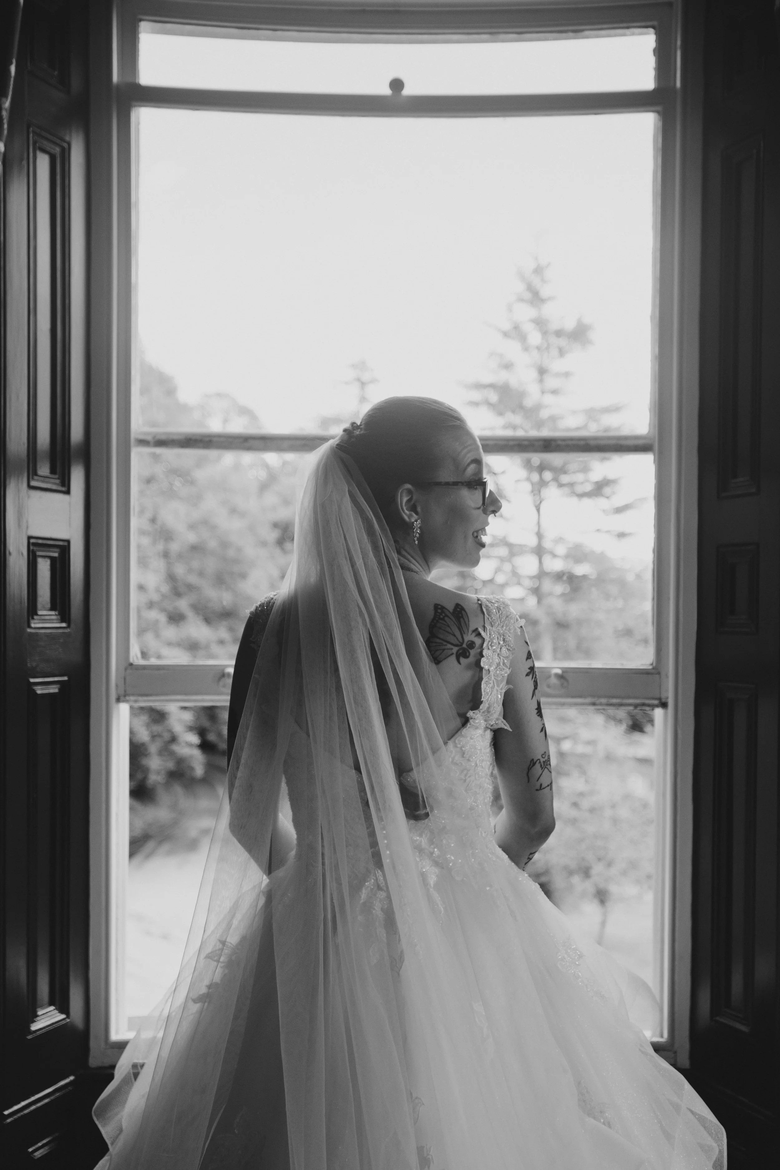 A bride wearing glasses and a wedding dress with a veil, standing in front of a large window with trees outside, smiling and looking to her left.