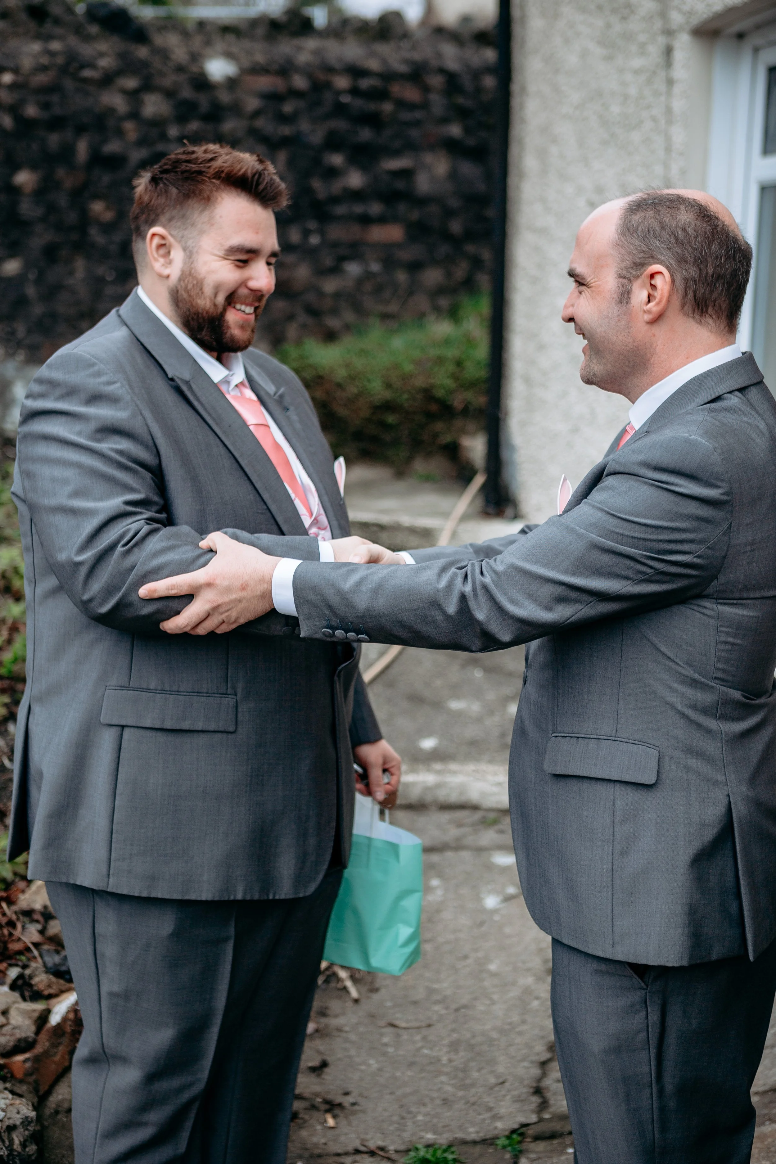 Two men in gray suits smiling and shaking hands outdoors, with one holding a gift bag, near a building with a stone wall.