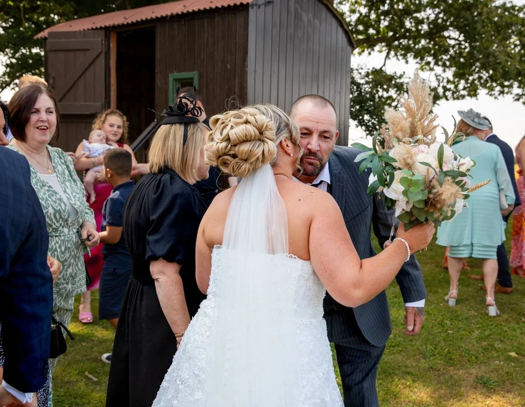 A bride and groom share an emotional moment during their wedding celebration outdoors, surrounded by family and friends near a rustic wooden building. The bride is holding a bouquet of flowers.