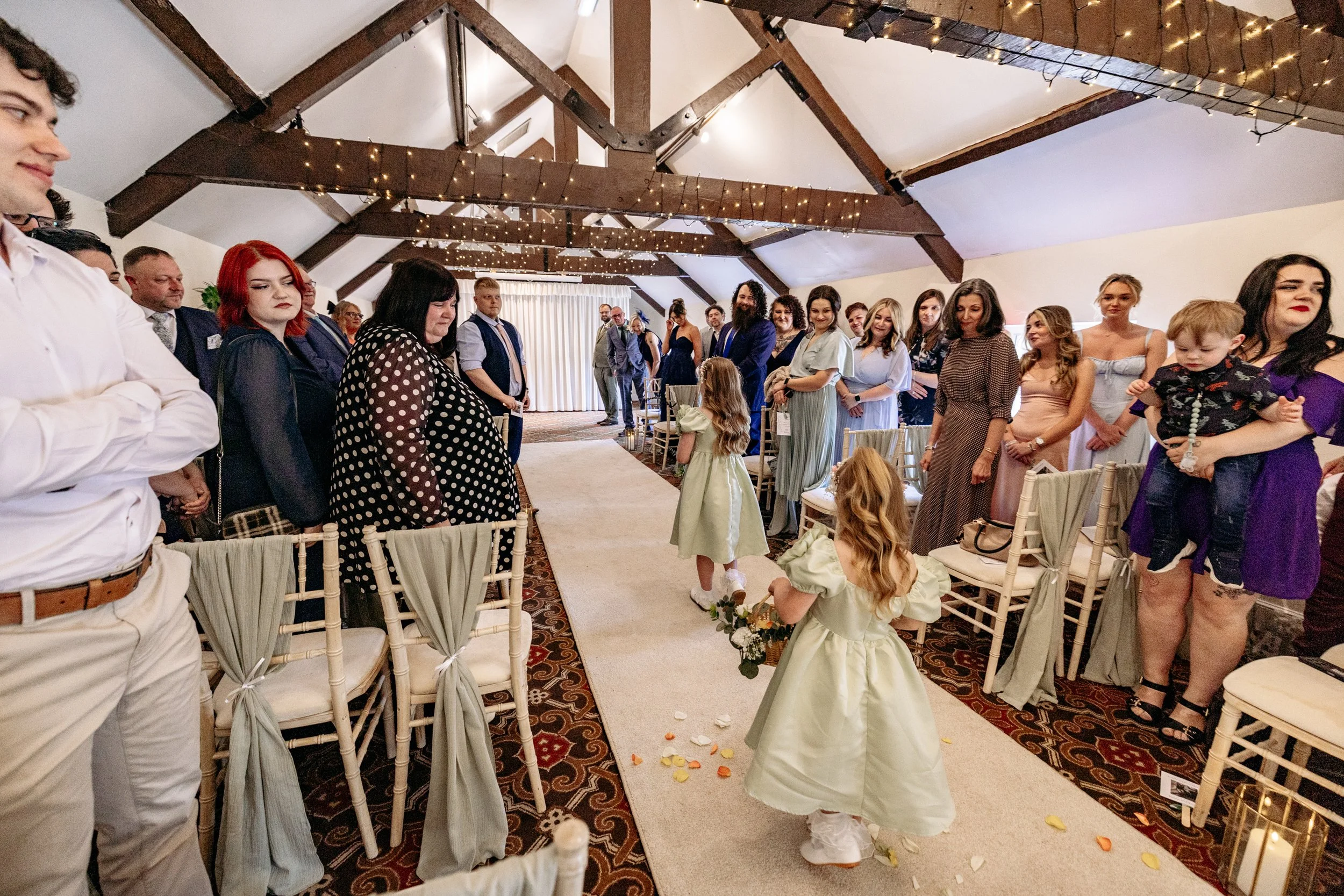 Two flower girls walking down the aisle at a wedding ceremony, with guests standing on either side in a decorated indoor venue with wooden beams and string lights.