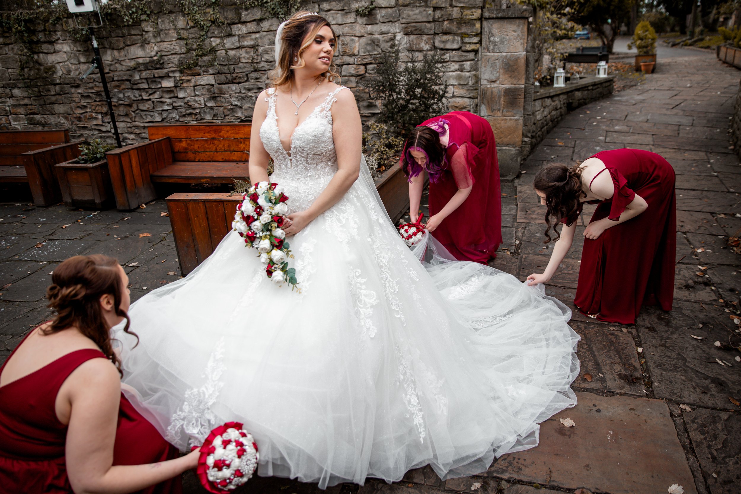 A bride in a white wedding gown stands outdoors while four women in burgundy dresses adjust her wedding dress. One woman holds a bouquet, another crouches, and the others are bending down to help with the dress train. The setting has stone paving and