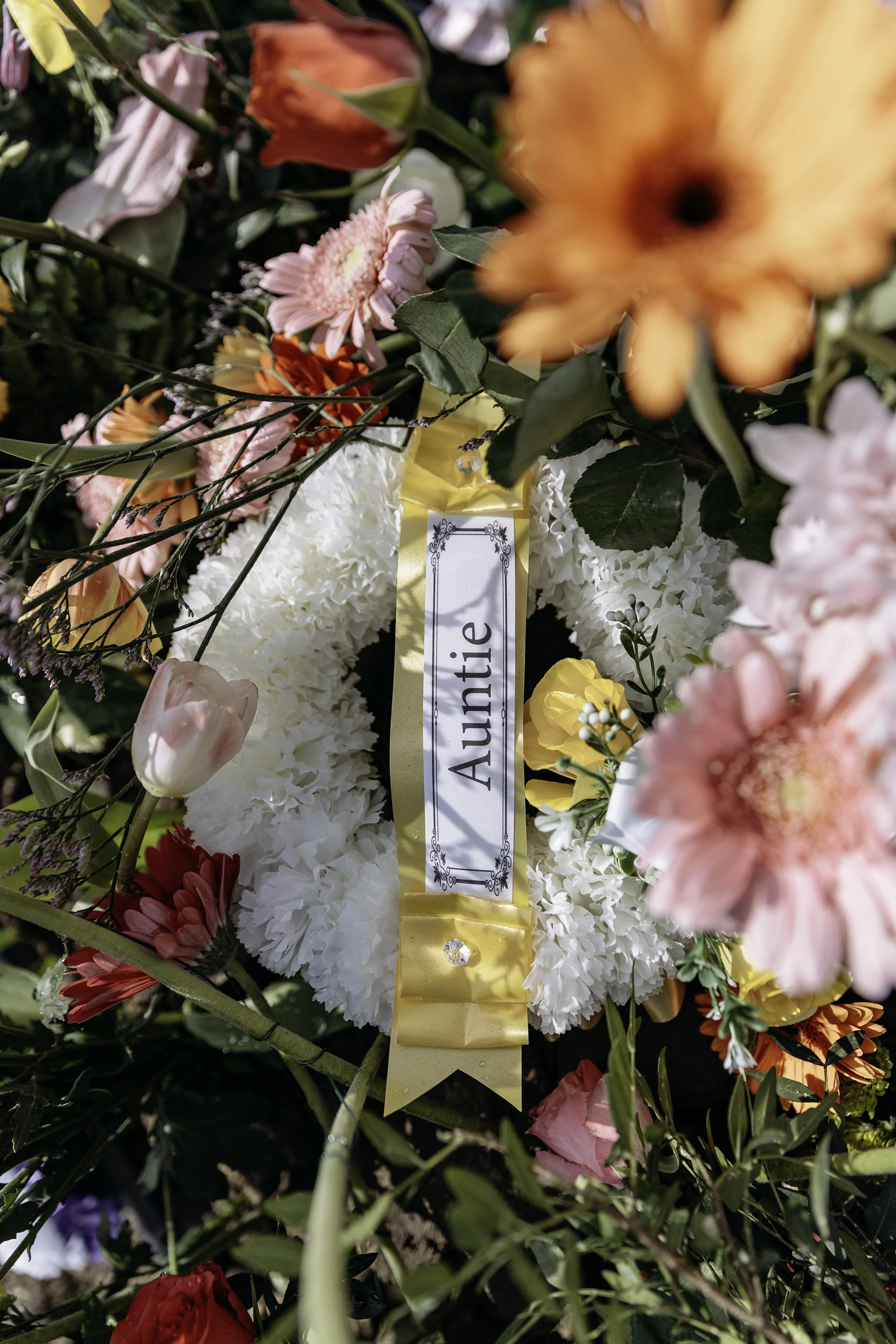 A floral arrangement with white, pink, orange, yellow, and red flowers surrounding a white wreath with a gold ribbon that has a label reading 'Auntie' in black text.