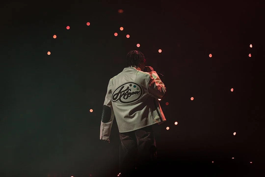 A person performing on stage with a microphone, wearing a jacket with a large logo on the back, surrounded by small red lights in a dark setting.