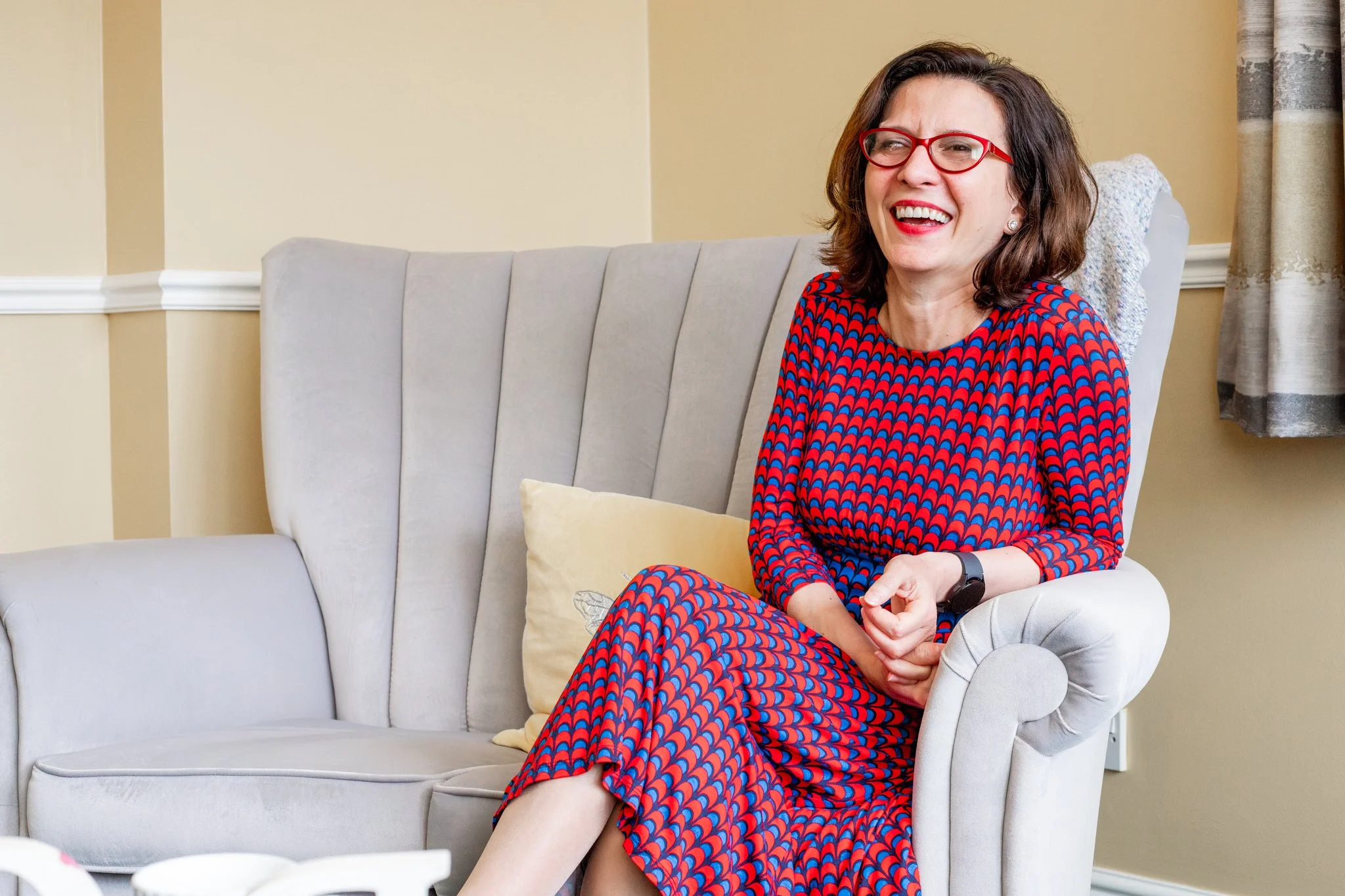 A woman with shoulder-length brown hair, red glasses, and a red-and-blue patterned dress sitting on a light gray upholstered armchair in a living room, smiling and laughing.