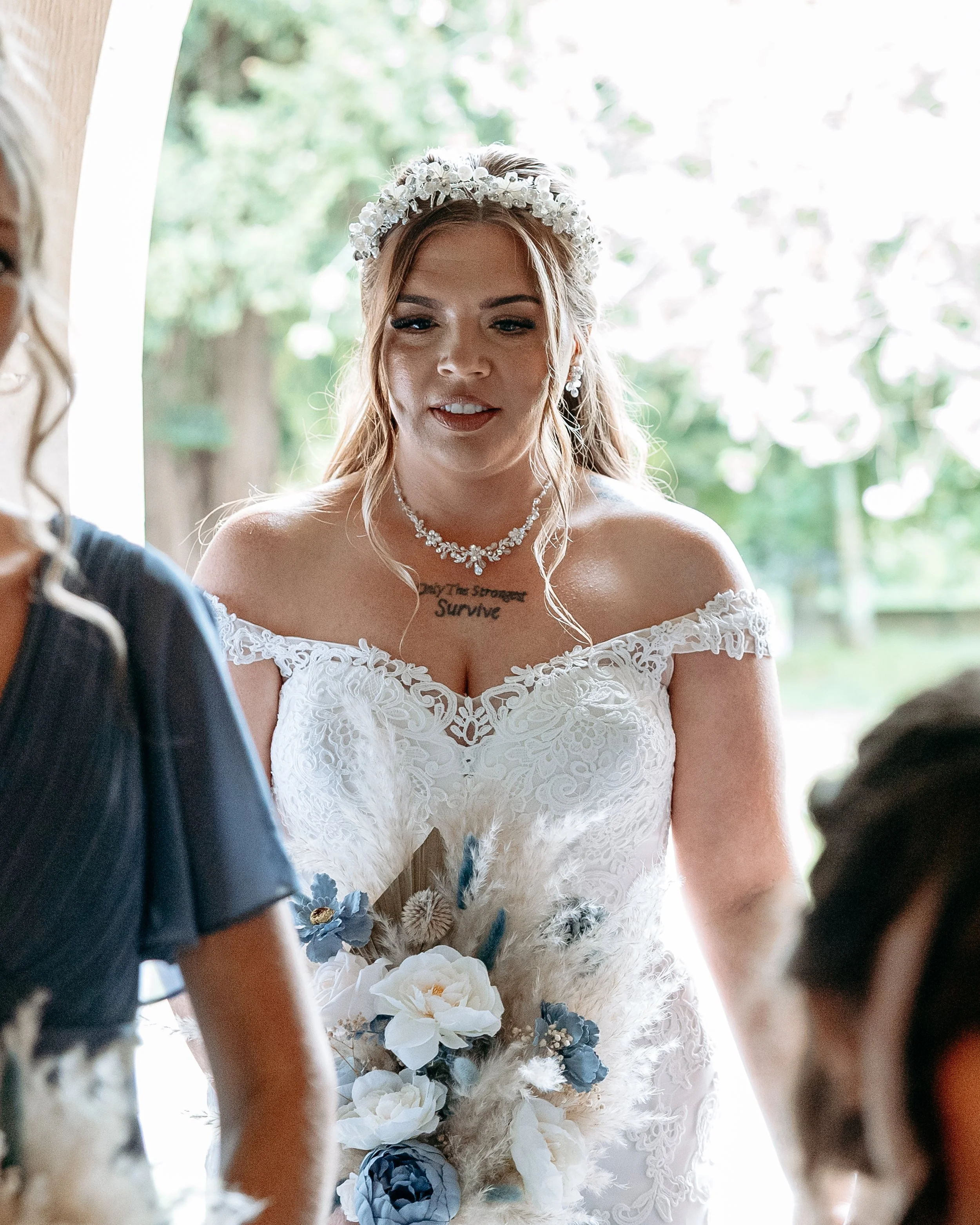A bride with a floral headband, off-the-shoulder lace wedding dress, and floral bouquet, standing indoors with greenery in the background.