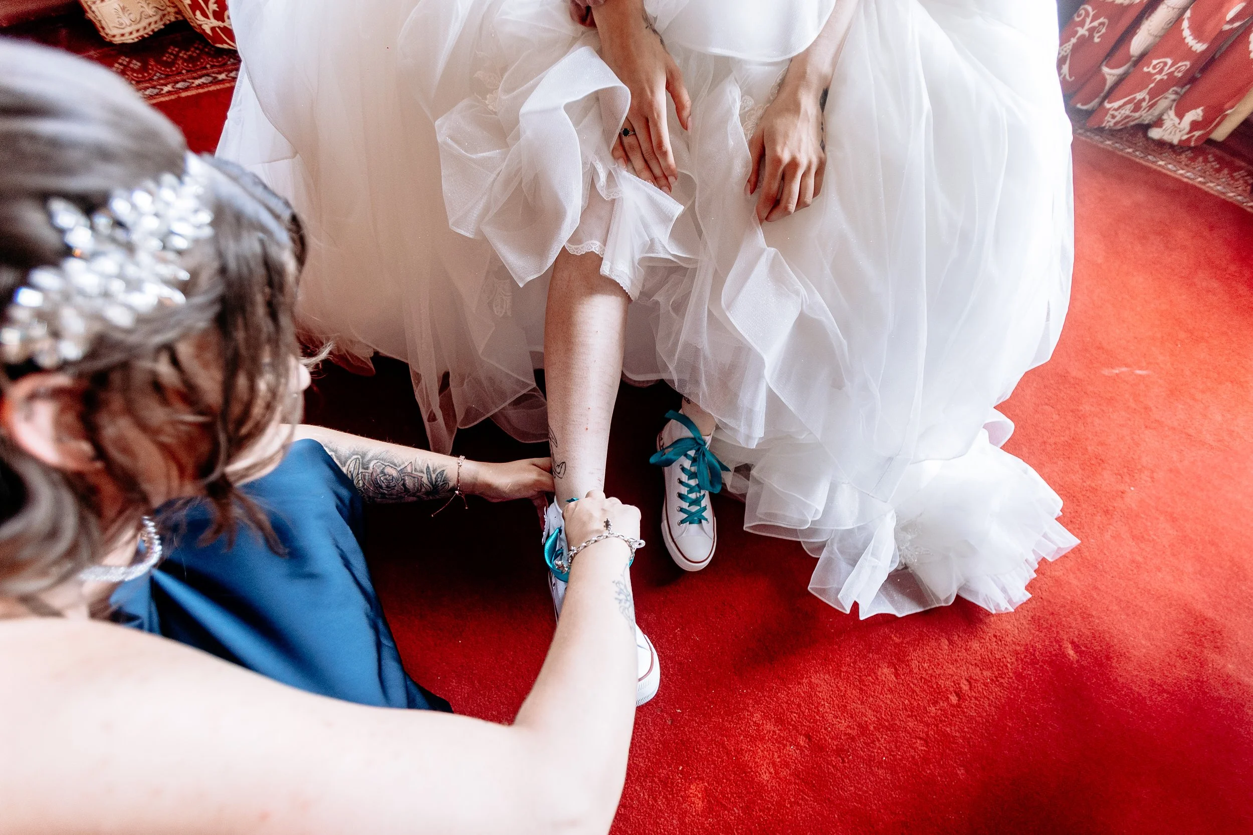 A bride in a white wedding dress with blue sneakers is having her leg adjusted by a woman in a blue dress. The scene is on a red carpet with decorative elements in the background.