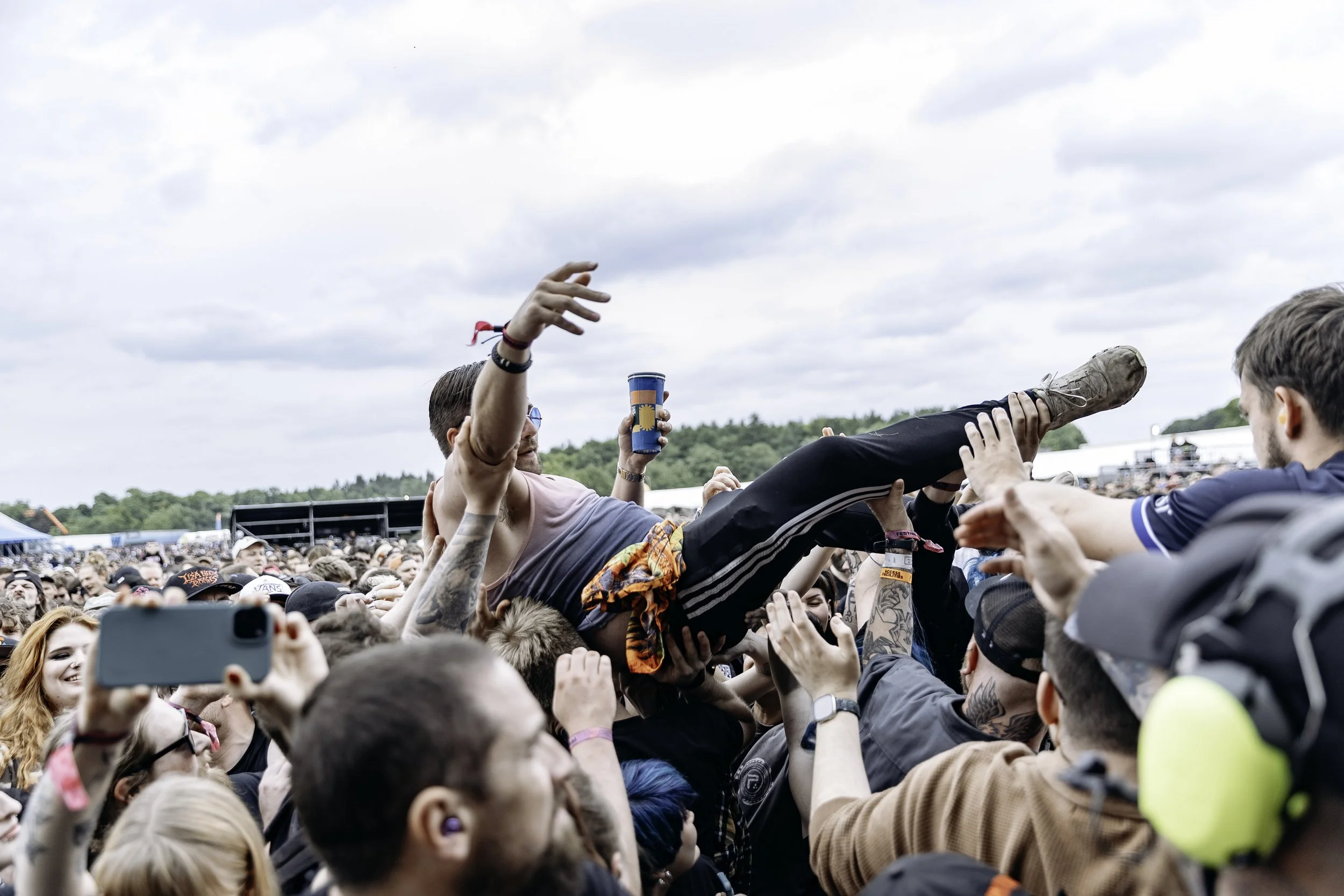 Crowd at a music festival crowd surfing in the outdoors during daytime, with cloudy sky and group of people supporting a person above the crowd.