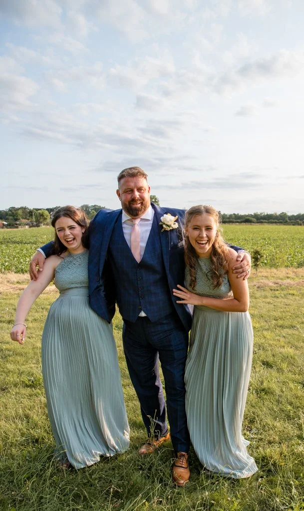 A man in a suit and two young girls in matching green dresses standing outdoors on a grassy field, smiling and posing for a photo.