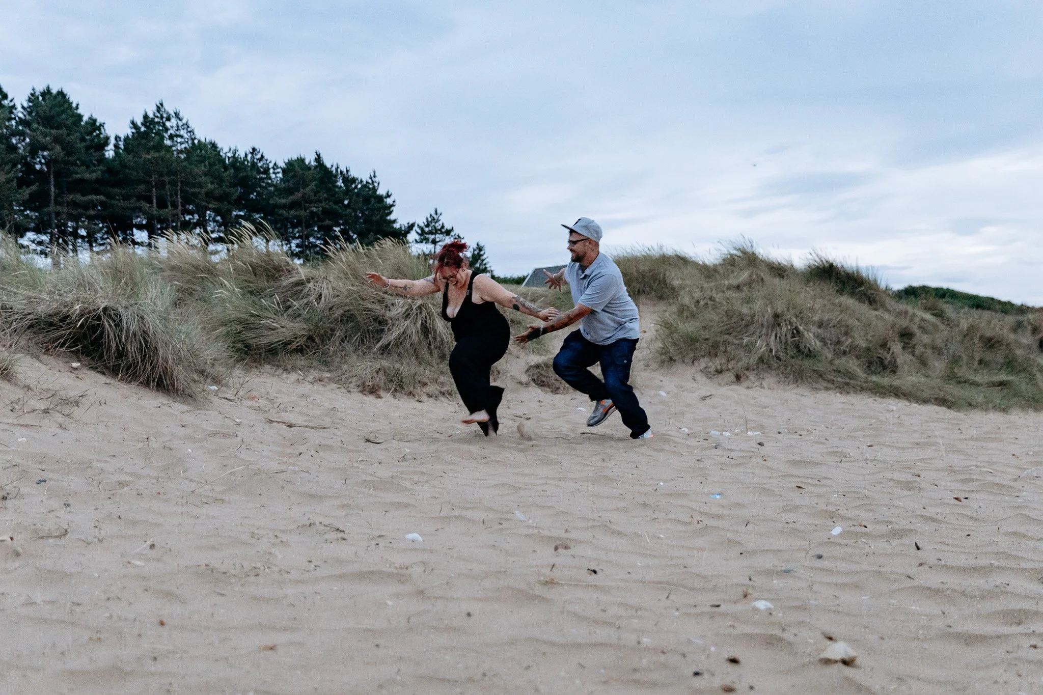 Two people, a woman in black dress and a man in jeans and gray shirt, are playing and running on a sandy beach near grassy dunes and trees.