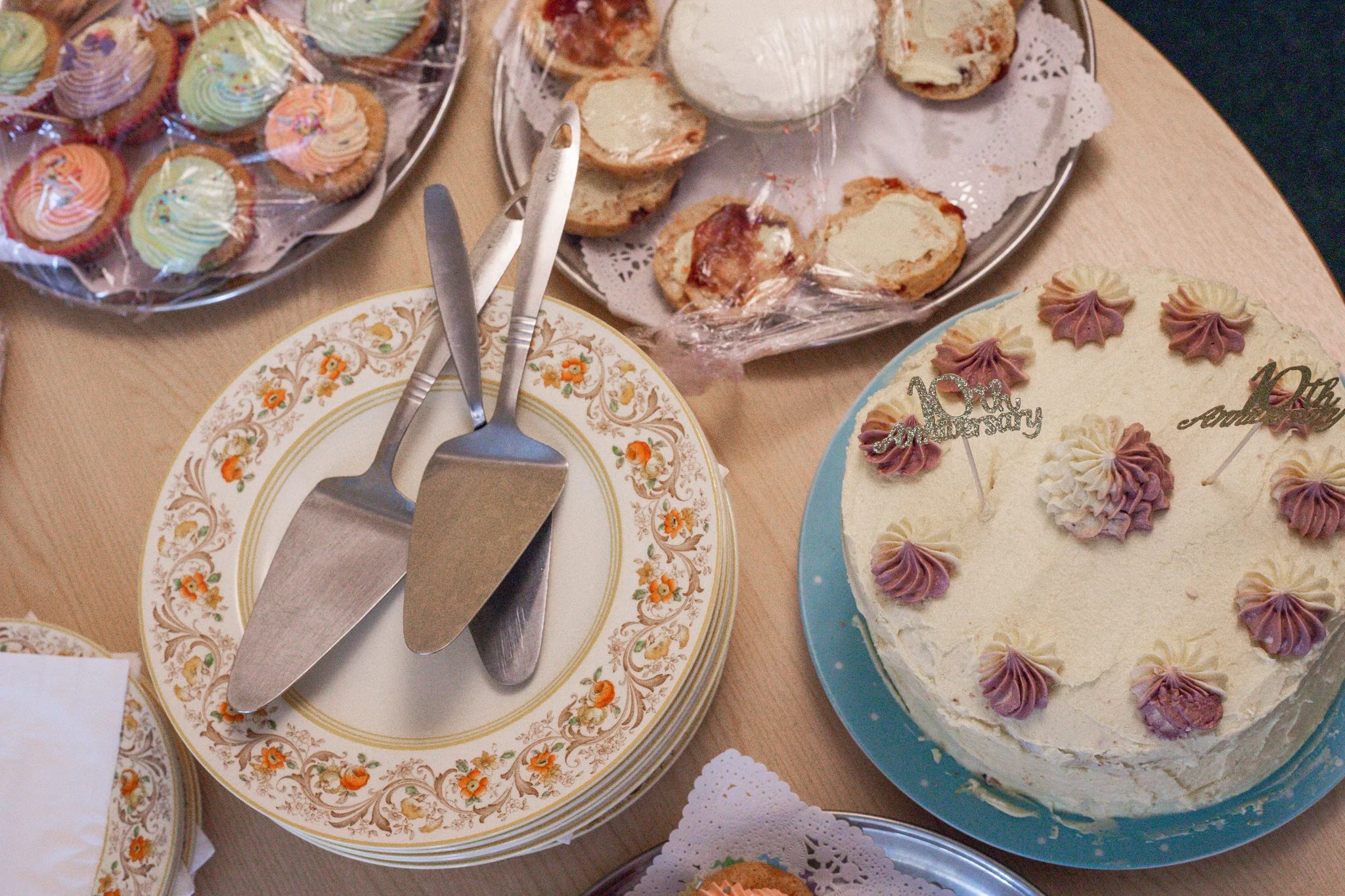 A table with a white birthday cake decorated with pastel purple and white buttercream flowers and 'Happy 40th Anniversary' cake toppers. Next to it, a stack of decorative plates with utensils on top. Surrounding the cake are plates of cupcakes with c