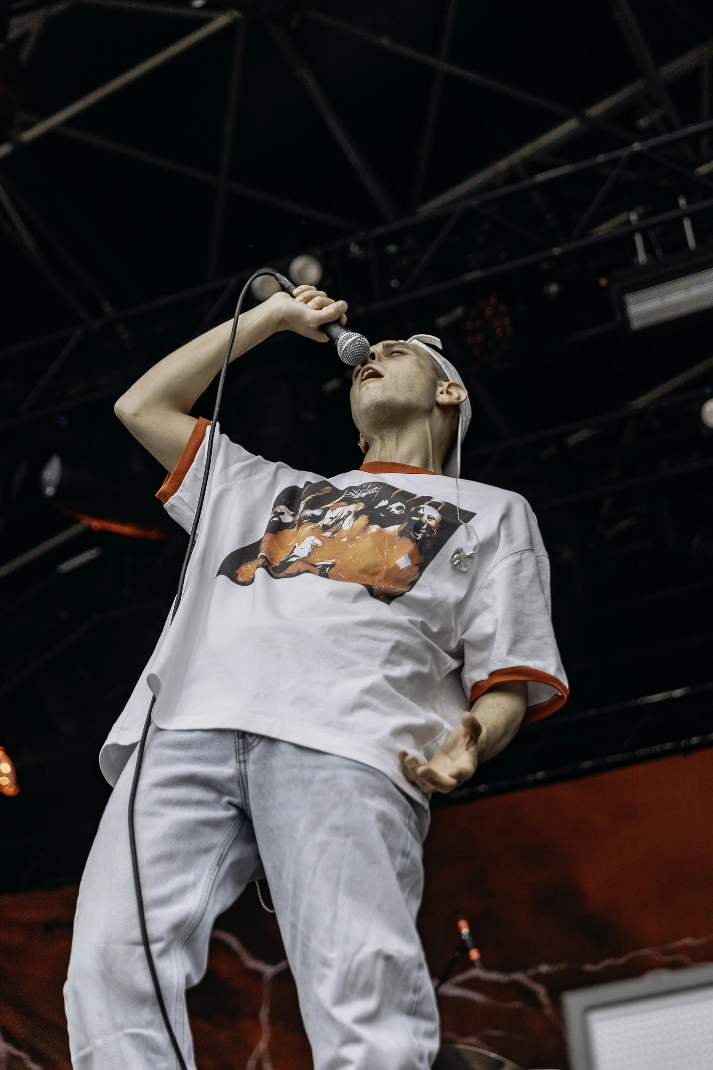 Male singer performing on stage, holding a microphone close to his mouth, with a black metal stage framework overhead and a red backdrop.